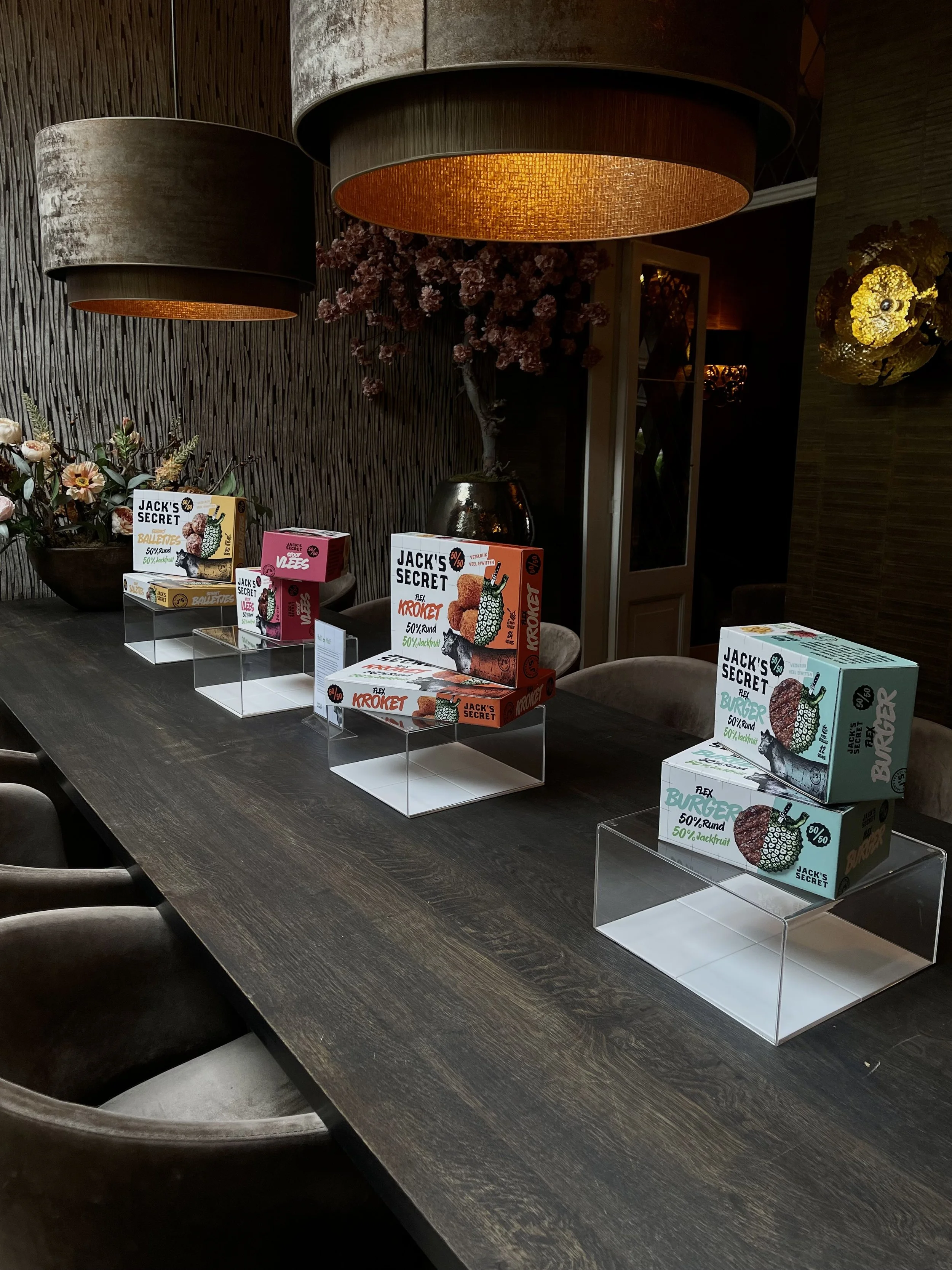 Mesa de madera con varias cajas de snack de la marca Jack's Secret y sillas alrededor, en un ambiente con iluminación cálida y decoraciones florales.