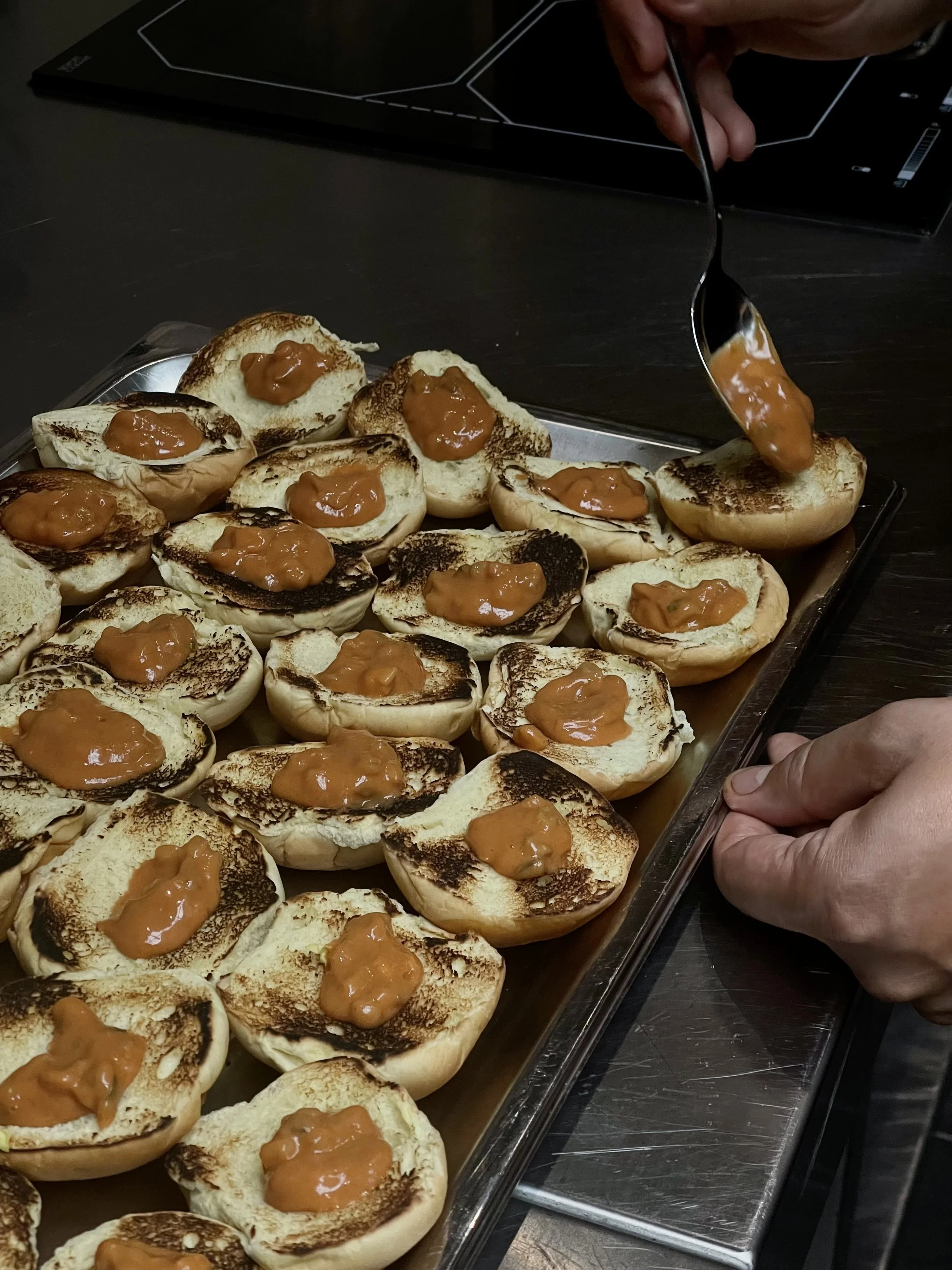 Unas personas están colocando mermelada de durazno en panes tostados, sobre una bandeja de metal. El fondo muestra una mesa de color oscuro y una caja negra con líneas blancas.