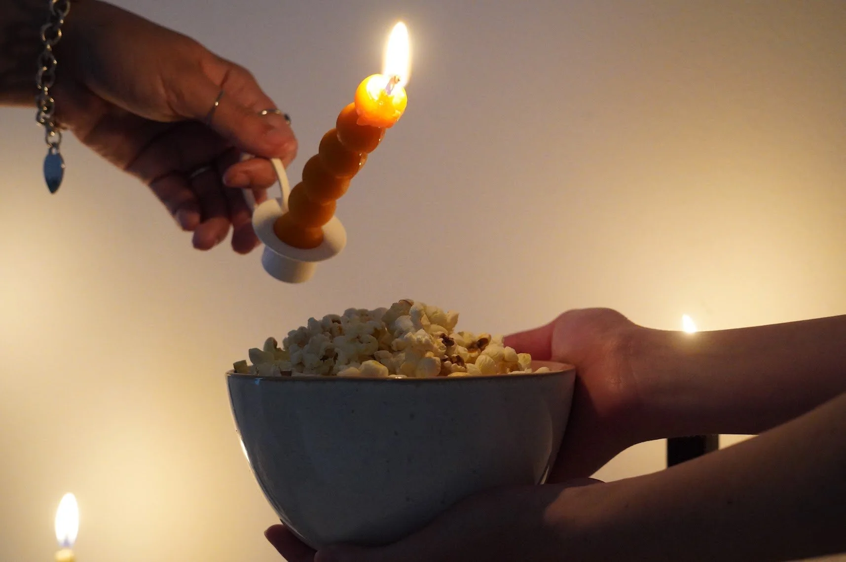 Dos manos, una con joyería, sostienen un tazón con palomitas de maíz. Una mano en la parte superior sostiene una vela de mantequilla, dirigida hacia las palomitas.