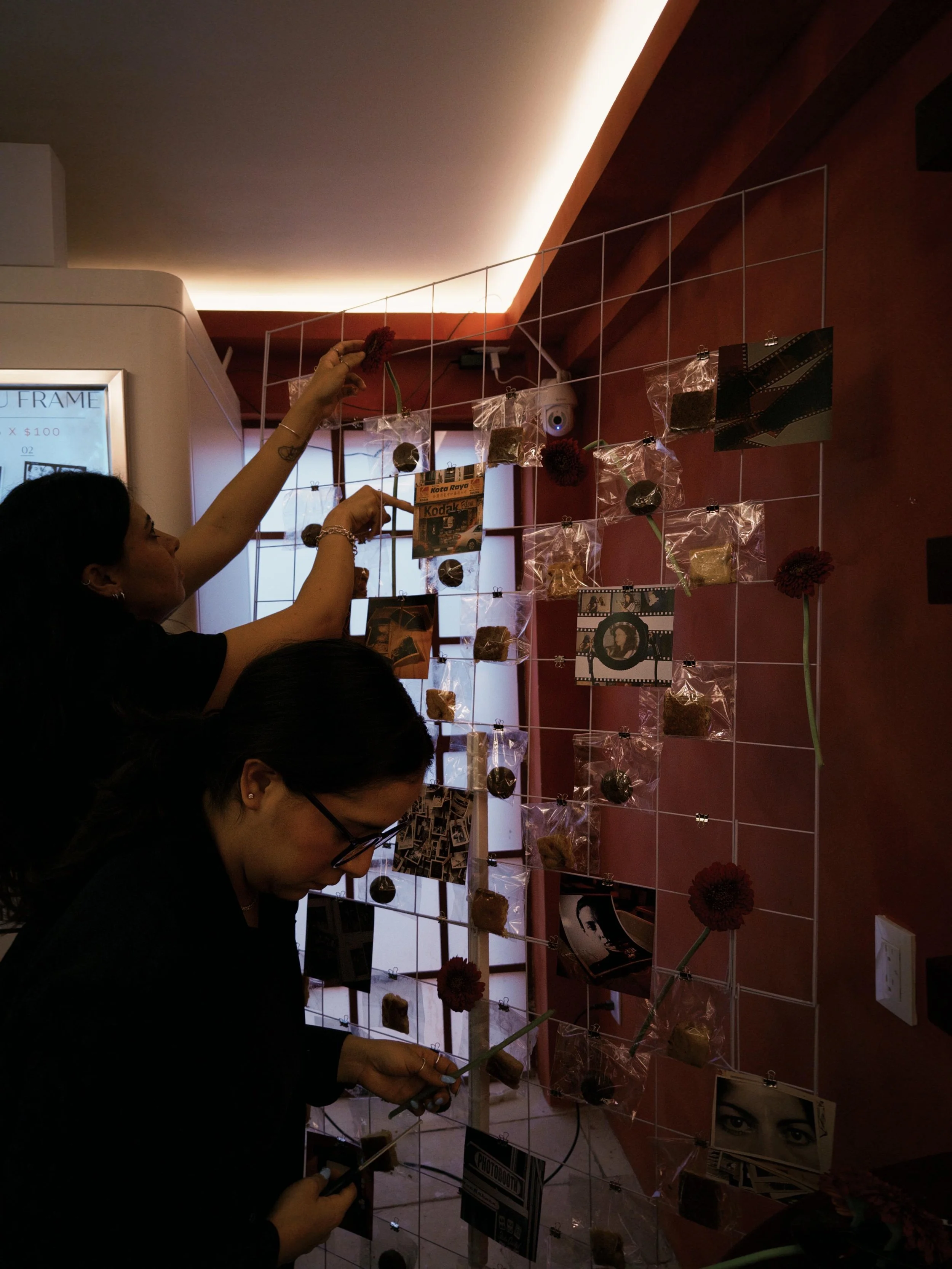 Varias personas decorando una pared con fotografías, posters y objetos pequeños, así como flores rojas en jarrones. Iluminación tenue en el fondo.