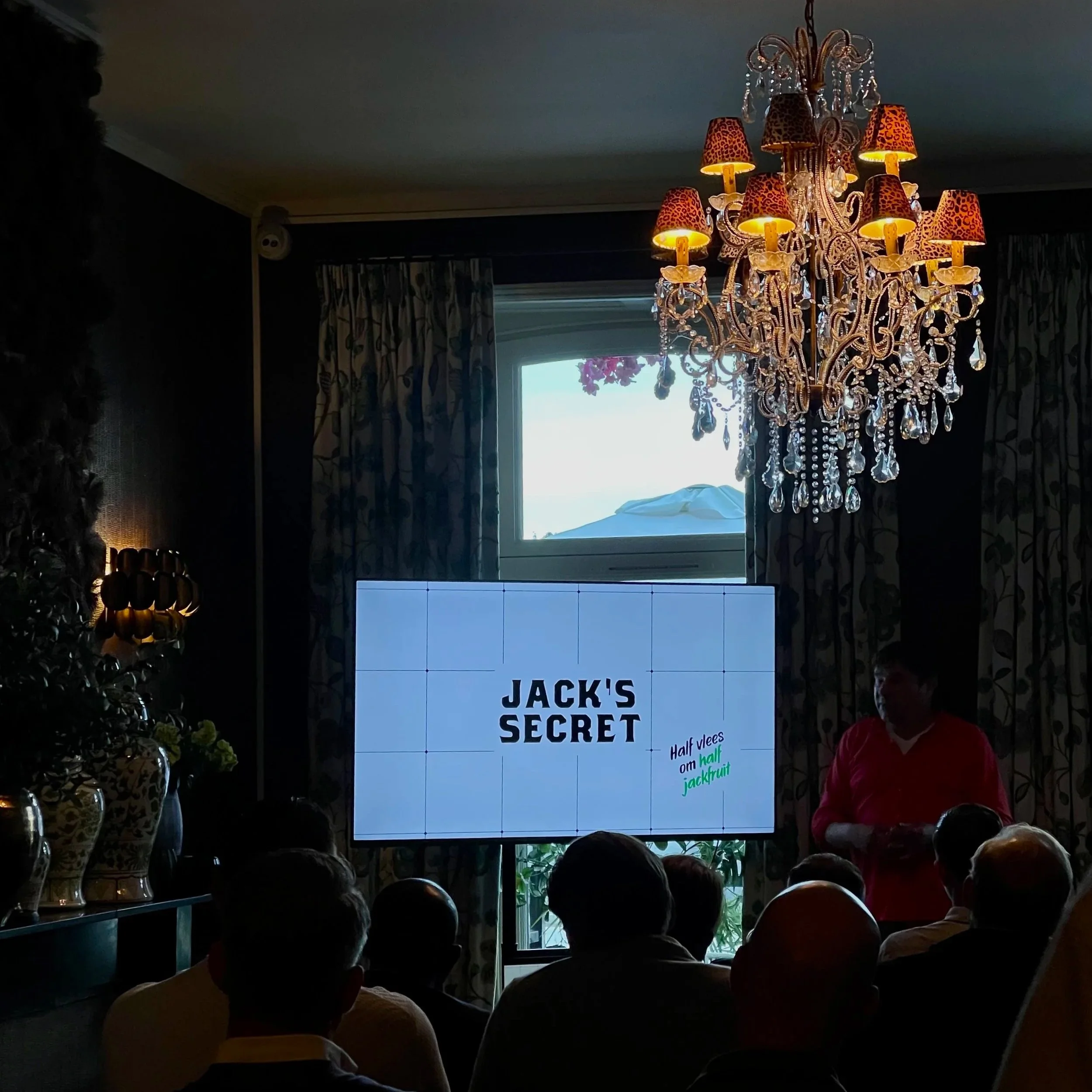 Pantalla digital que muestra el texto 'JACK'S SECRET' en letras negras, con fondo blanco, en una reunión o evento con múltiples personas y un candelabro decorativo con pantallas con estampado de leopardo y cristales colgantes.