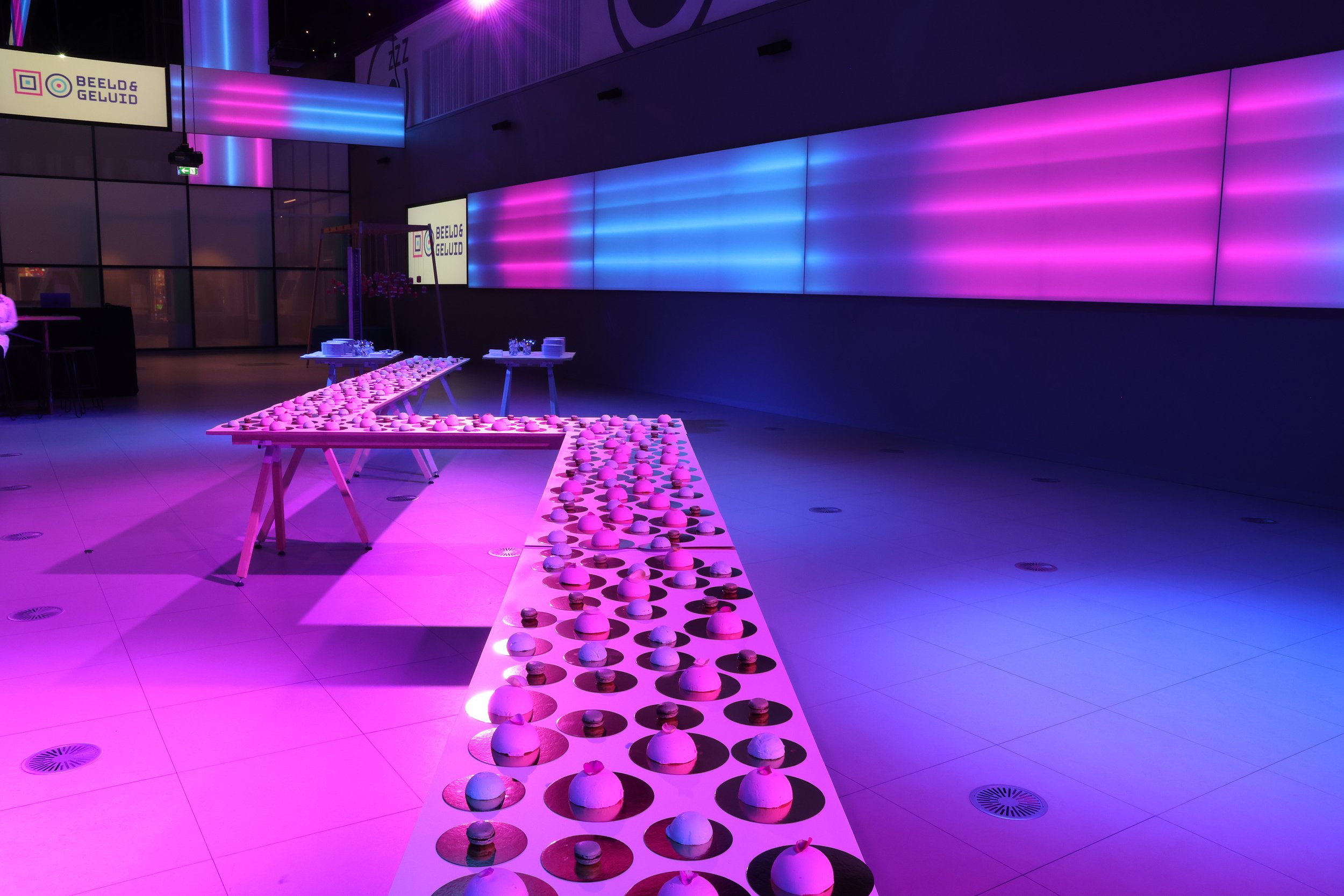 Sala con paredes decoradas con luces de neón en tonos morados y azules, mesas con comida y objetos decorativos en forma de cúpula, en un ambiente de evento moderno y vibrante.