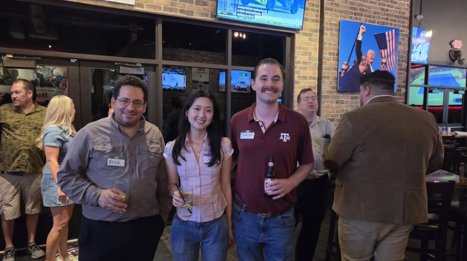 Three people pose at a casual restaurant or sports bar, with televisions and patrons in the background.