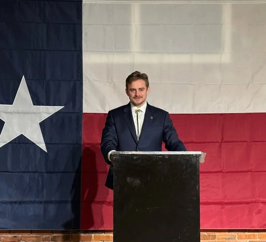 Man in a suit speaking at a podium in front of a Texas flag