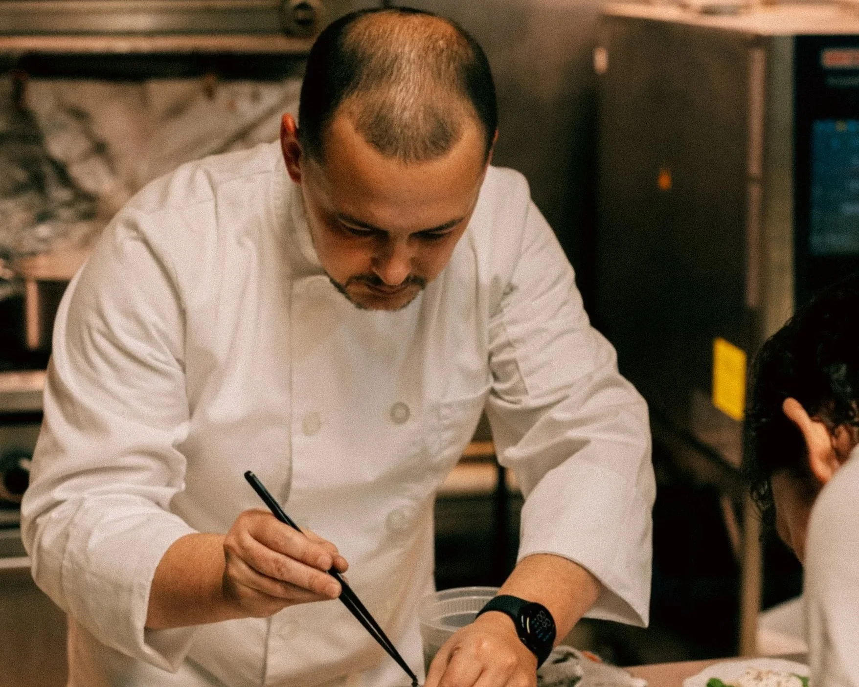 A chef in a white uniform and wristwatch writing in a notebook in a professional kitchen.