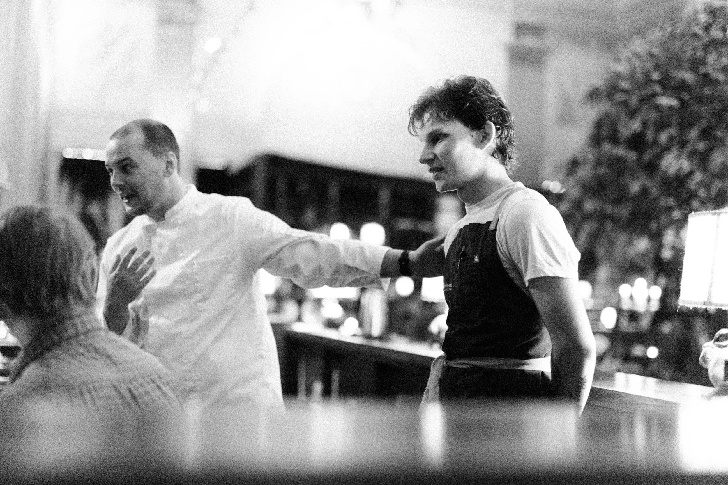 Two young men are standing close together in a social setting, engaging in conversation with smiles and gestures, while surrounded by other people in a warmly lit, indoor environment.