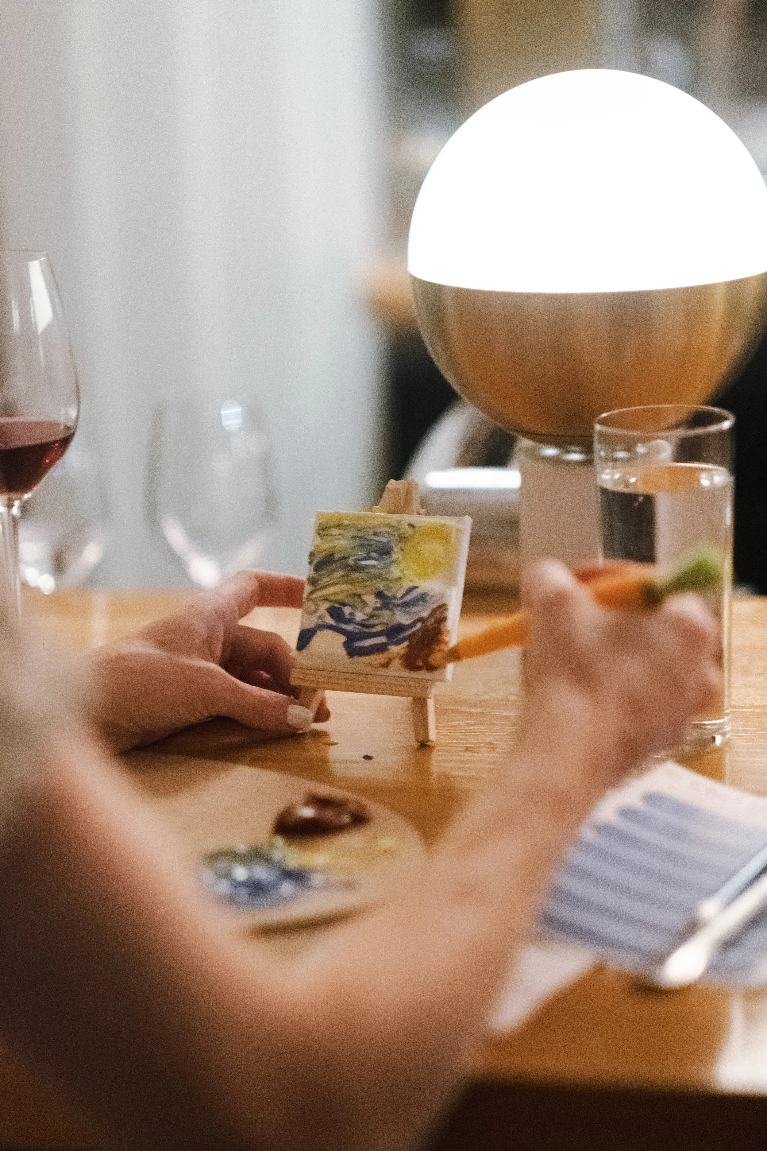 Person holding a piece of artwork with a painted landscape scene, sitting at a table with wine glasses and a table lamp in the background.