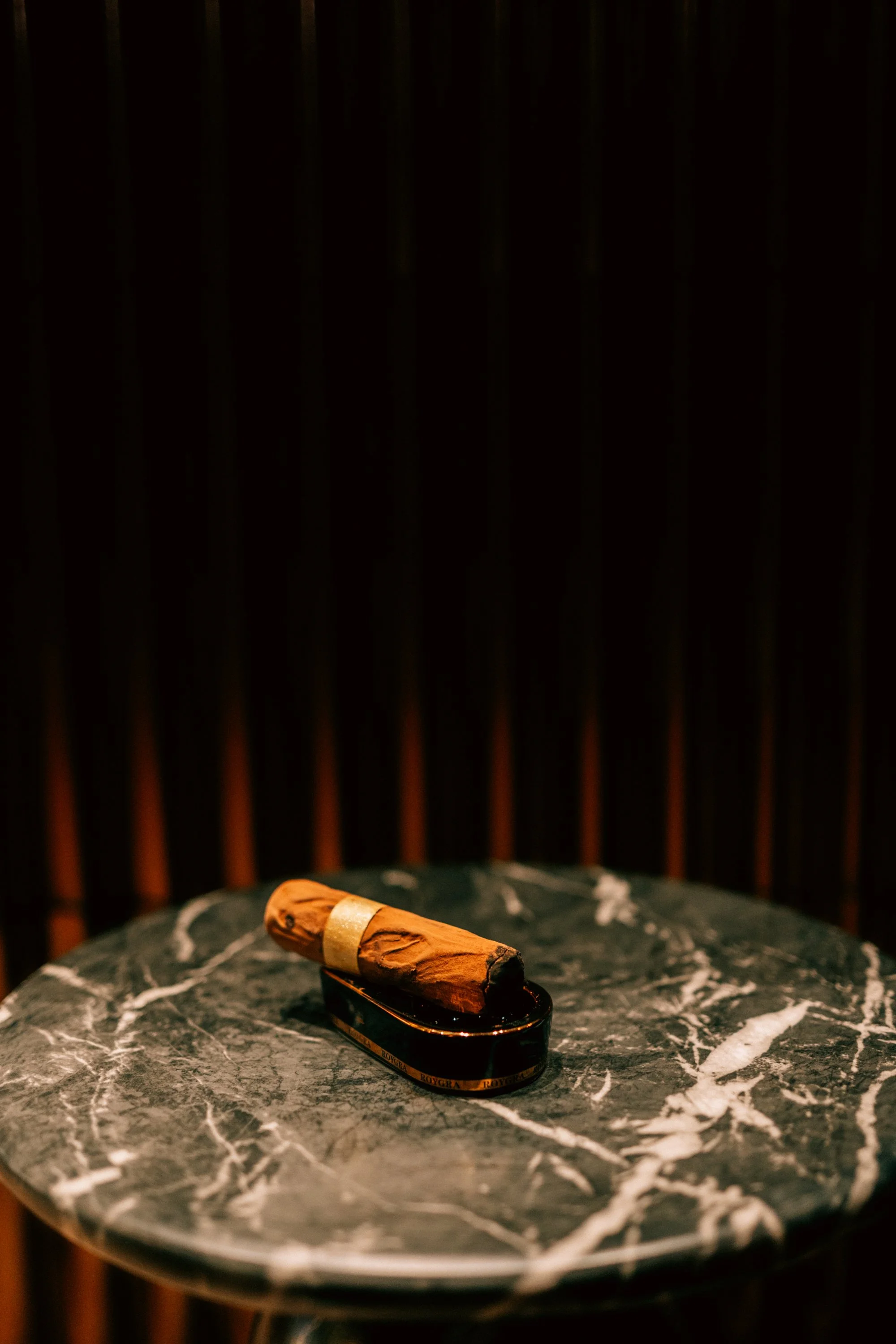 A lit cigar on a black and gold ashtray, placed on a round, dark marble table with white veining.