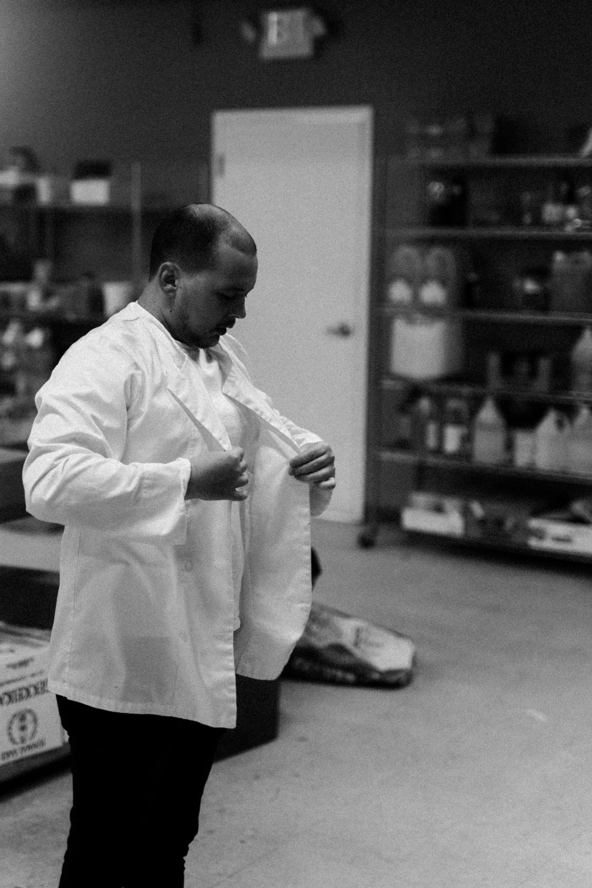 A person with short hair wearing a white coat, standing indoors, looking down, with shelves of various items in the background.