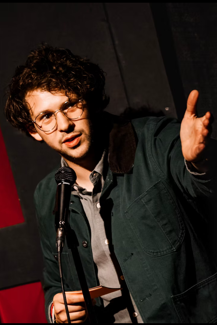 Young man with curly hair and glasses speaking into a microphone, gesturing with his right hand, on a stage with dark background.