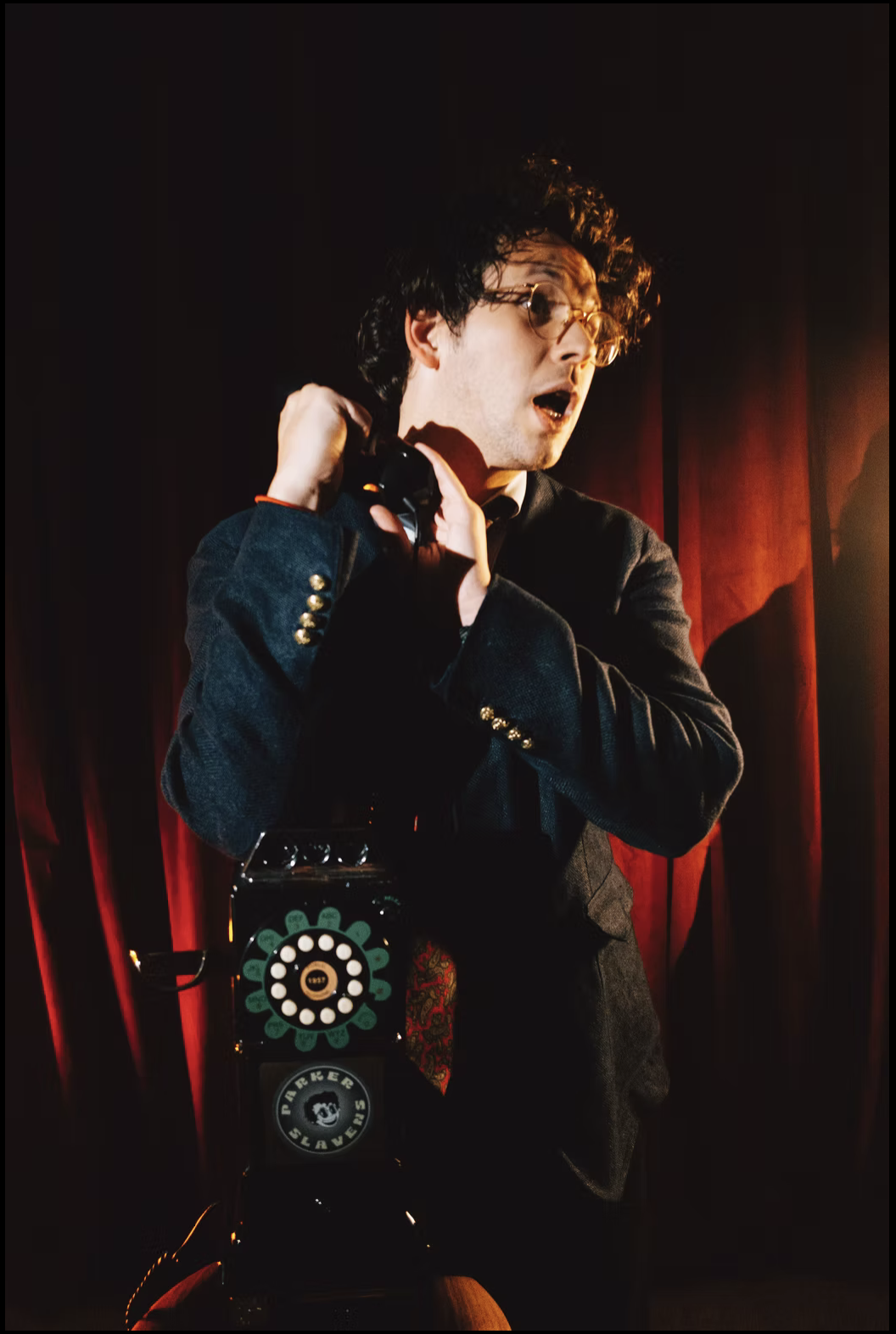 A man with curly dark hair and glasses, dressed in a dark jacket with gold buttons, holds a vintage black rotary phone with a colorful flower design at the base. He appears surprised or shocked, standing in front of red curtains with warm lighting.