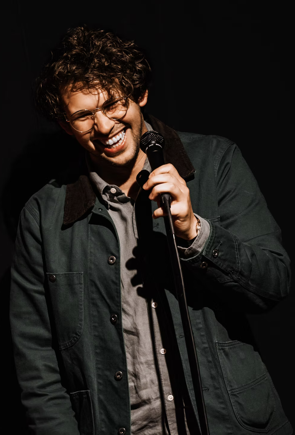 Young man with curly hair and glasses holding a microphone, laughing on stage.