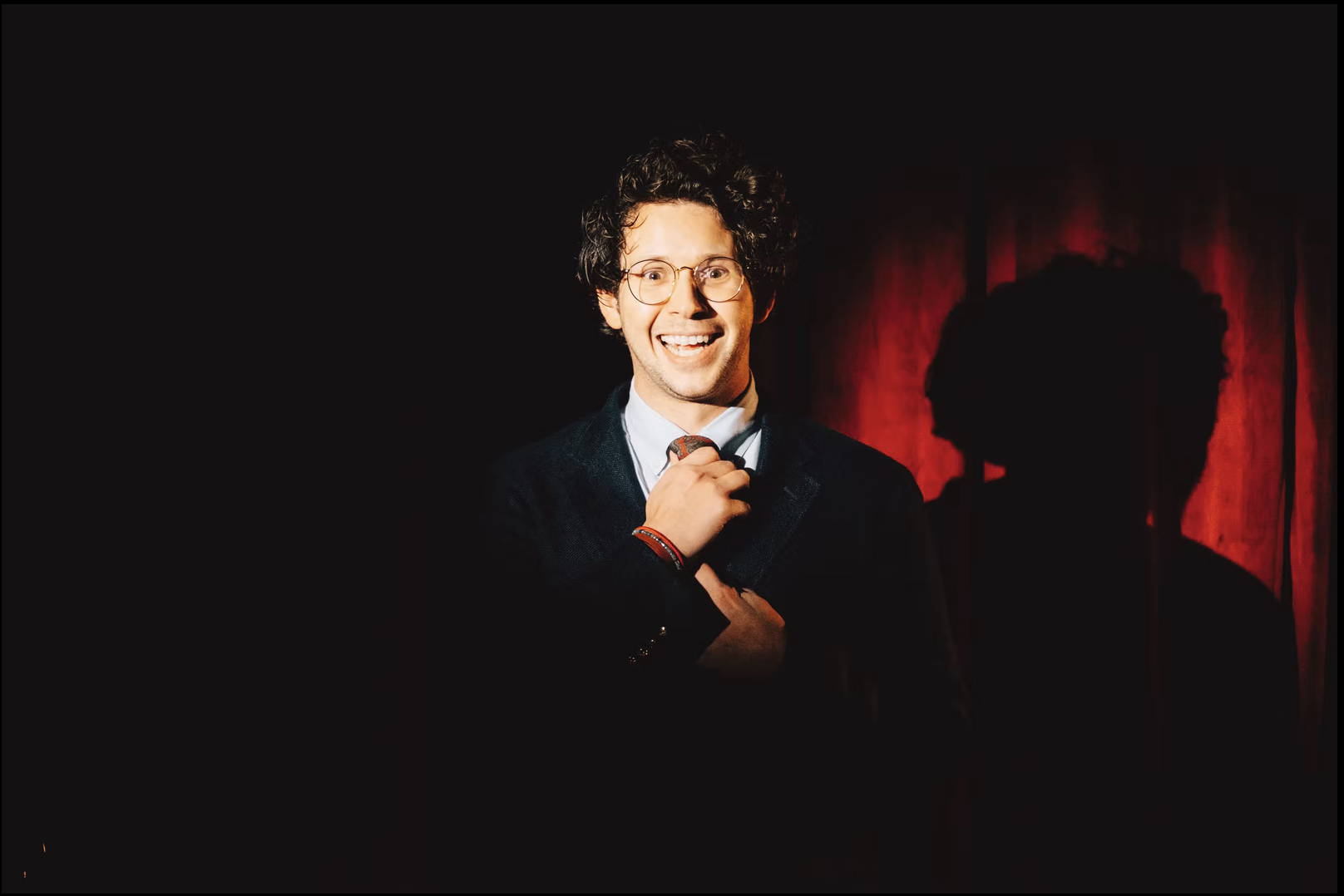 A man with curly dark hair and glasses smiling, dressed in a dark suit, holding his tie, standing in front of red curtains with shadowy profile behind him.