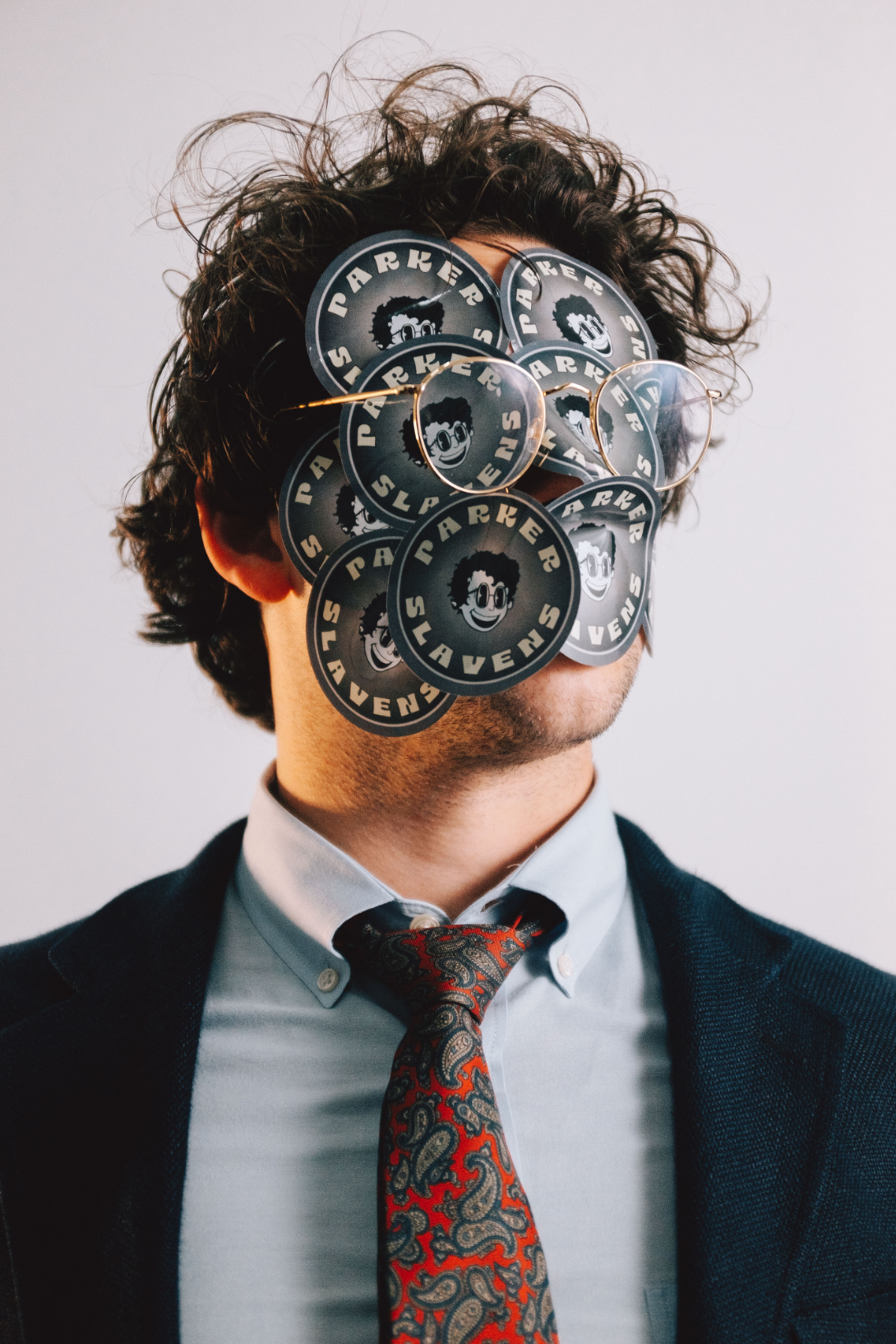 Person with curly hair and glasses, wearing a suit and tie, faces to the right, with multiple stickers covering their face. The stickers have a cartoon face with sunglasses and the words 'Parkers Slavens'.