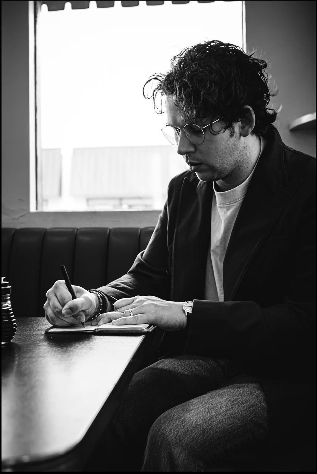 A black and white photo of a man with curly hair wearing glasses and a watch, sitting at a table, writing in a notebook with a pen, with a water bottle on the table, inside a cafe near a window.