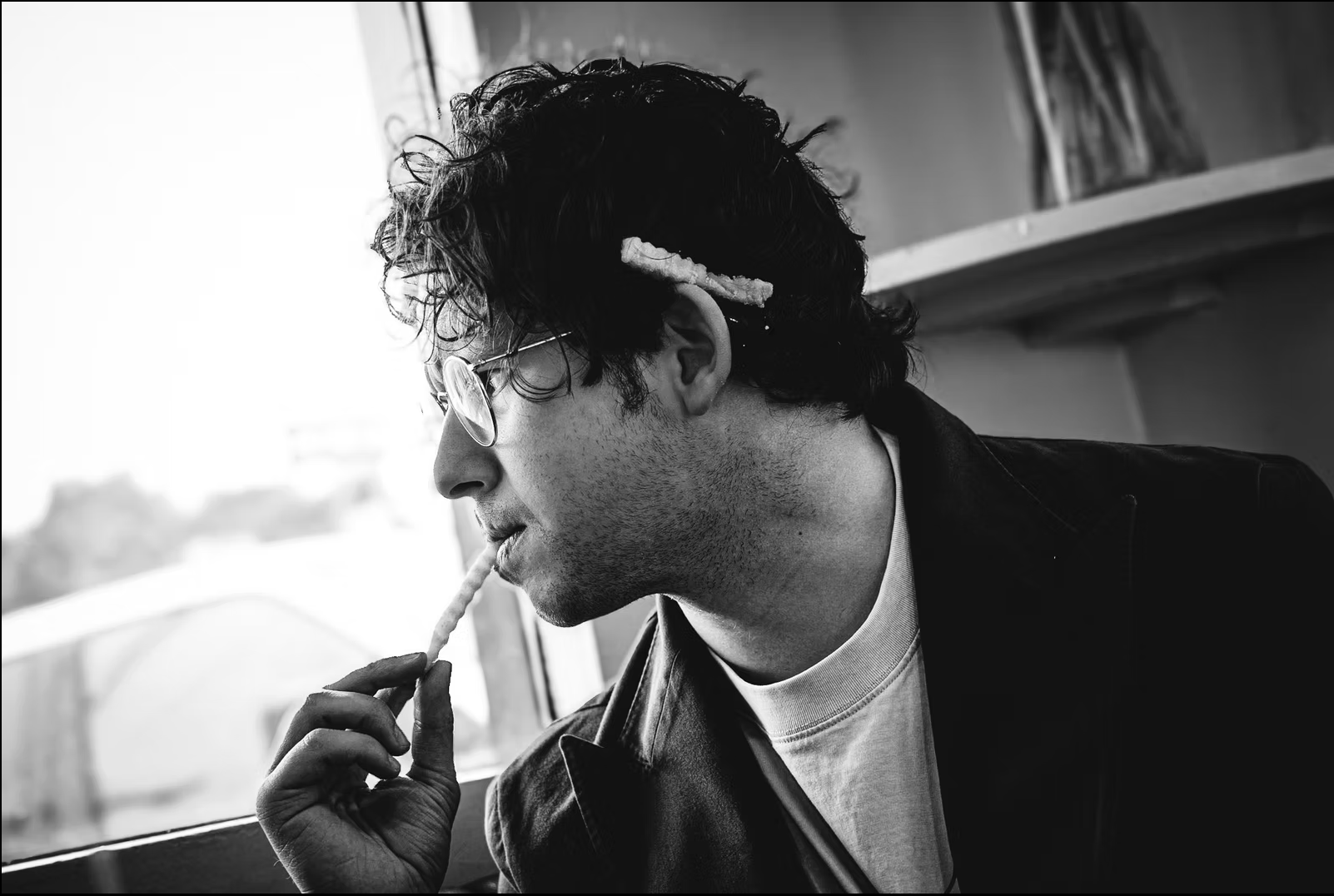 A black and white photo of a man with curly hair, wearing glasses, casually holding a cigarette near his lips, sitting in front of a window.