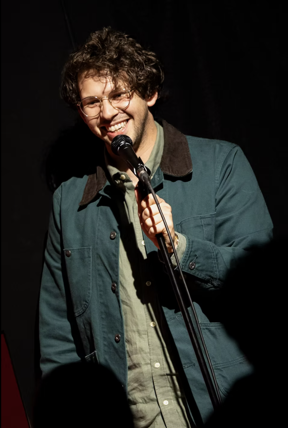 A man with curly hair, glasses, and a beard holding a microphone while smiling on stage.
