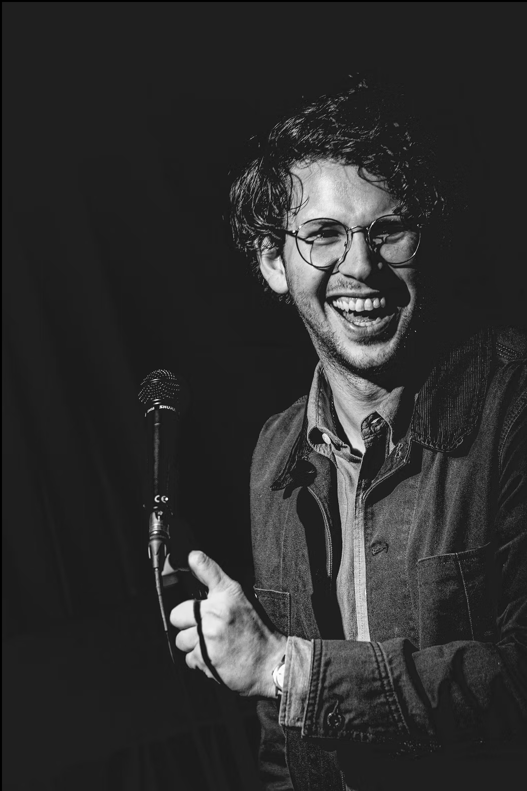 A young man with curly hair and glasses smiling and holding a microphone, wearing a denim jacket, in a black and white photo.