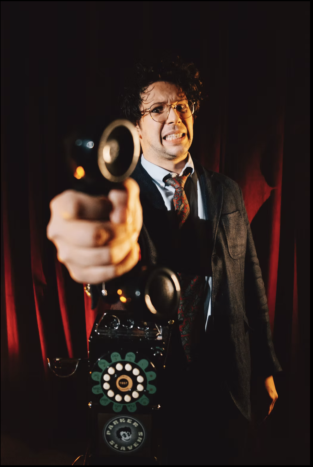 A man with glasses and messy hair points a vintage black rotary phone directly at the camera. He is dressed in a dark suit with a patterned tie and is standing in front of red velvet curtains, with a serious or intense facial expression.