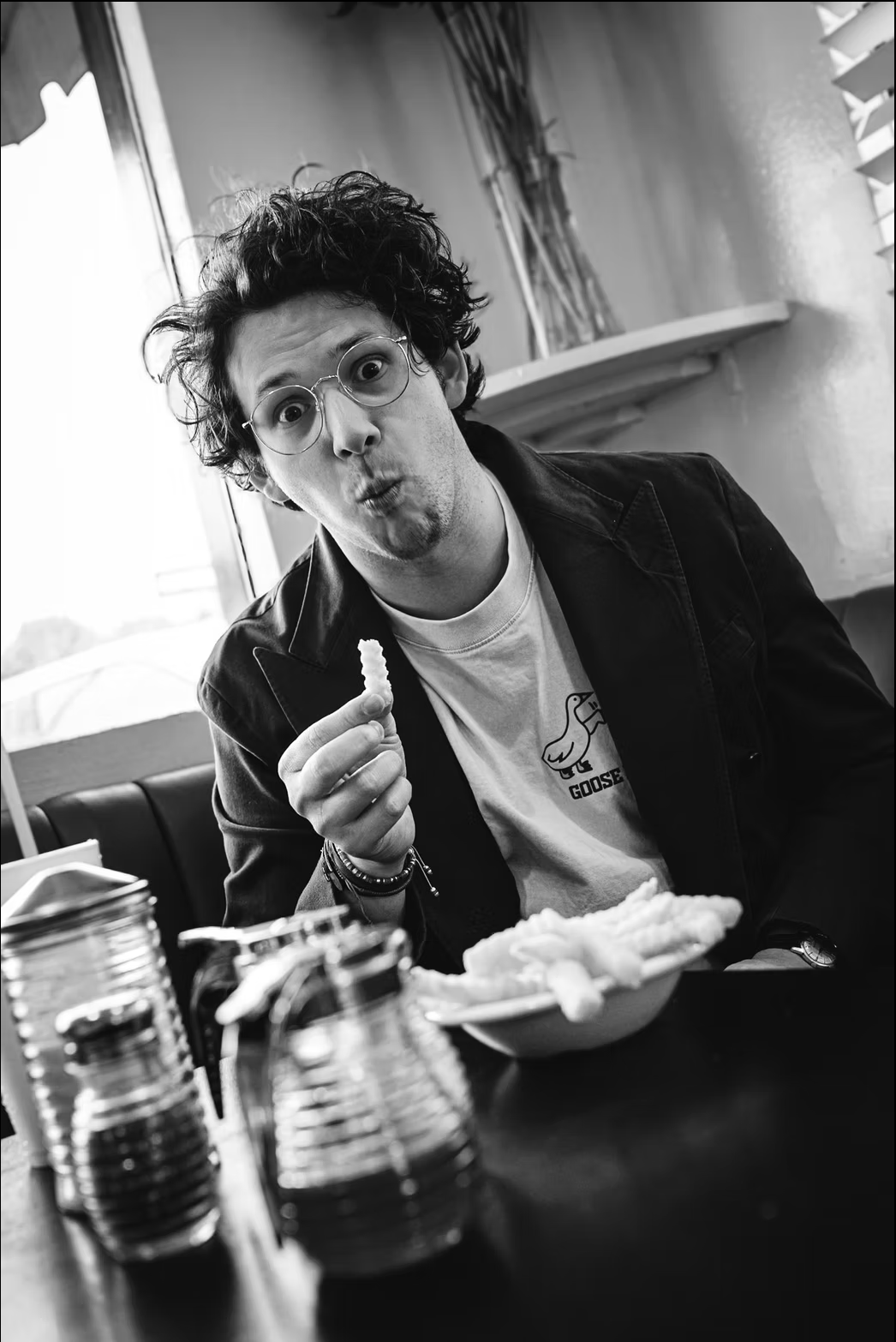 A young man with curly hair, glasses, and a light beard, making a surprised face while holding a chicken nugget in a restaurant. He's wearing a white T-shirt with a duck illustration and the word 'GOOSE' and a dark jacket. There are condiments on the