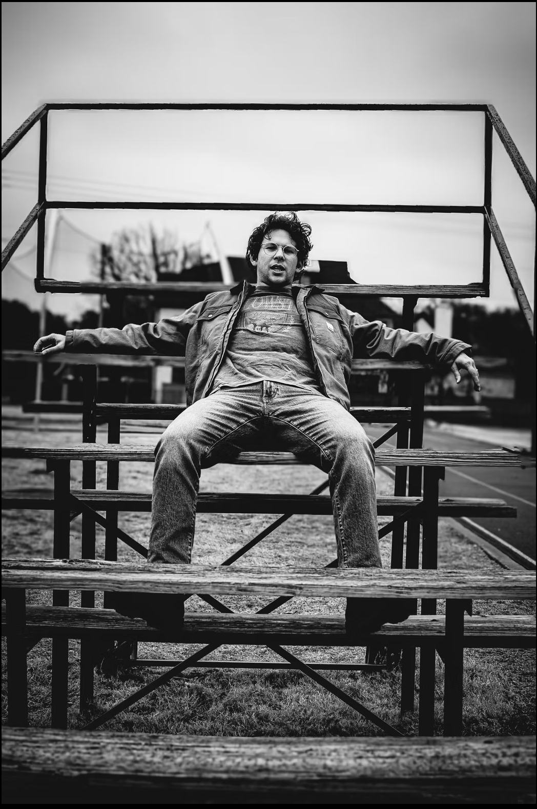 A black and white photo of a man with curly hair and glasses sitting on a wooden bleacher with his arms outstretched, outdoors on a cloudy day.