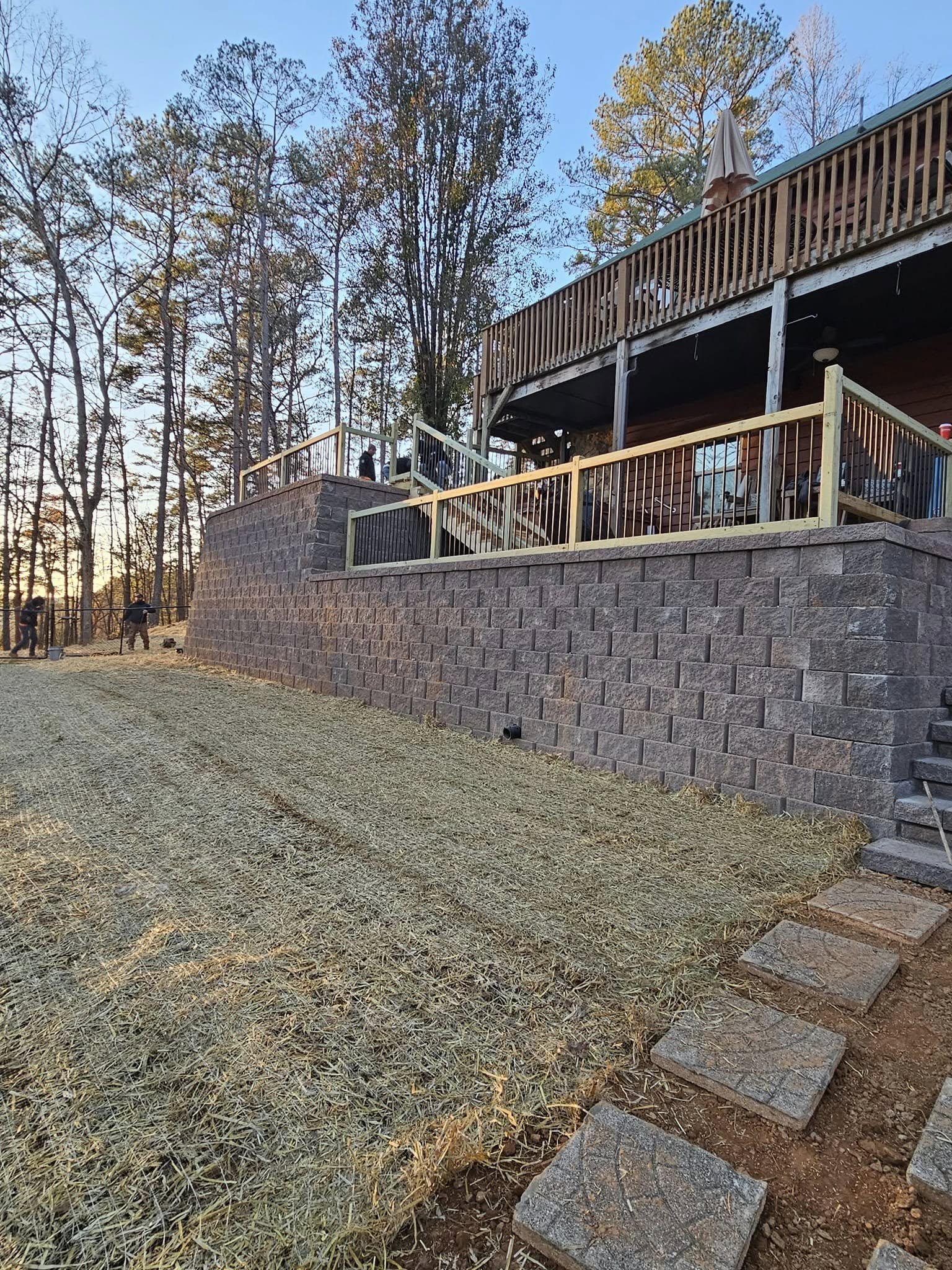 A backyard with a multi-level wooden deck, an unfinished railing, and a stone retaining wall. There are trees with fall foliage and a clear sky in the background.