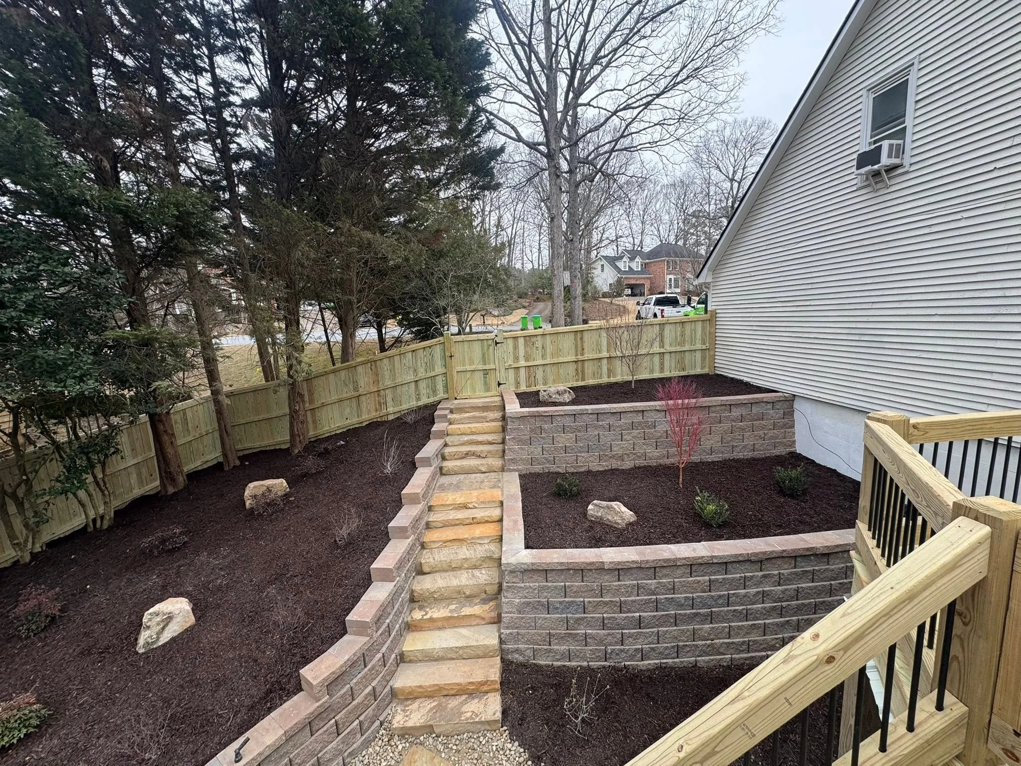 A backyard with a newly constructed raised garden bed with brick walls and steps, a wooden fence, and trees in the background. A house with a white exterior and window air conditioning unit is visible on the right.