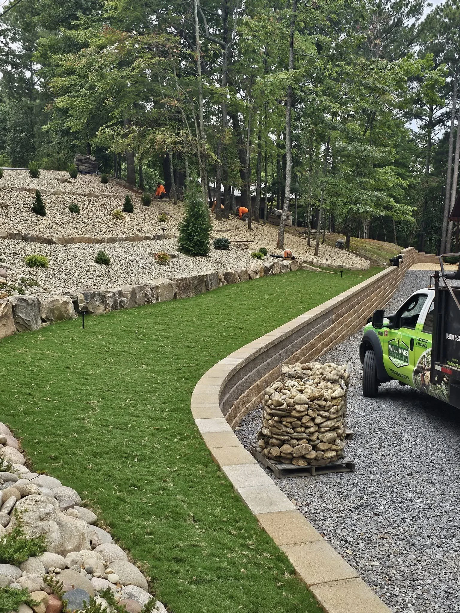 Landscaping work in progress on a sloped yard with rocks, small trees, and mulch, with workers and a truck present.