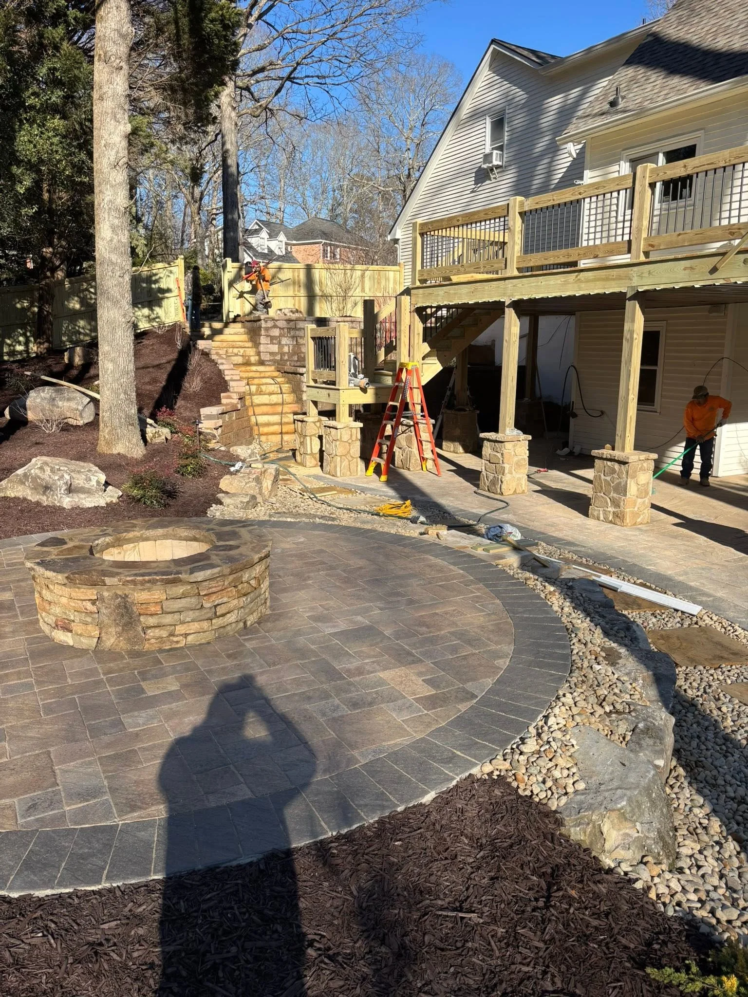 A backyard patio with a stone fire pit, a paved area, and a newly built wooden deck with stairs and railings. Construction workers are working on the deck, and the scene includes trees and a house with siding.