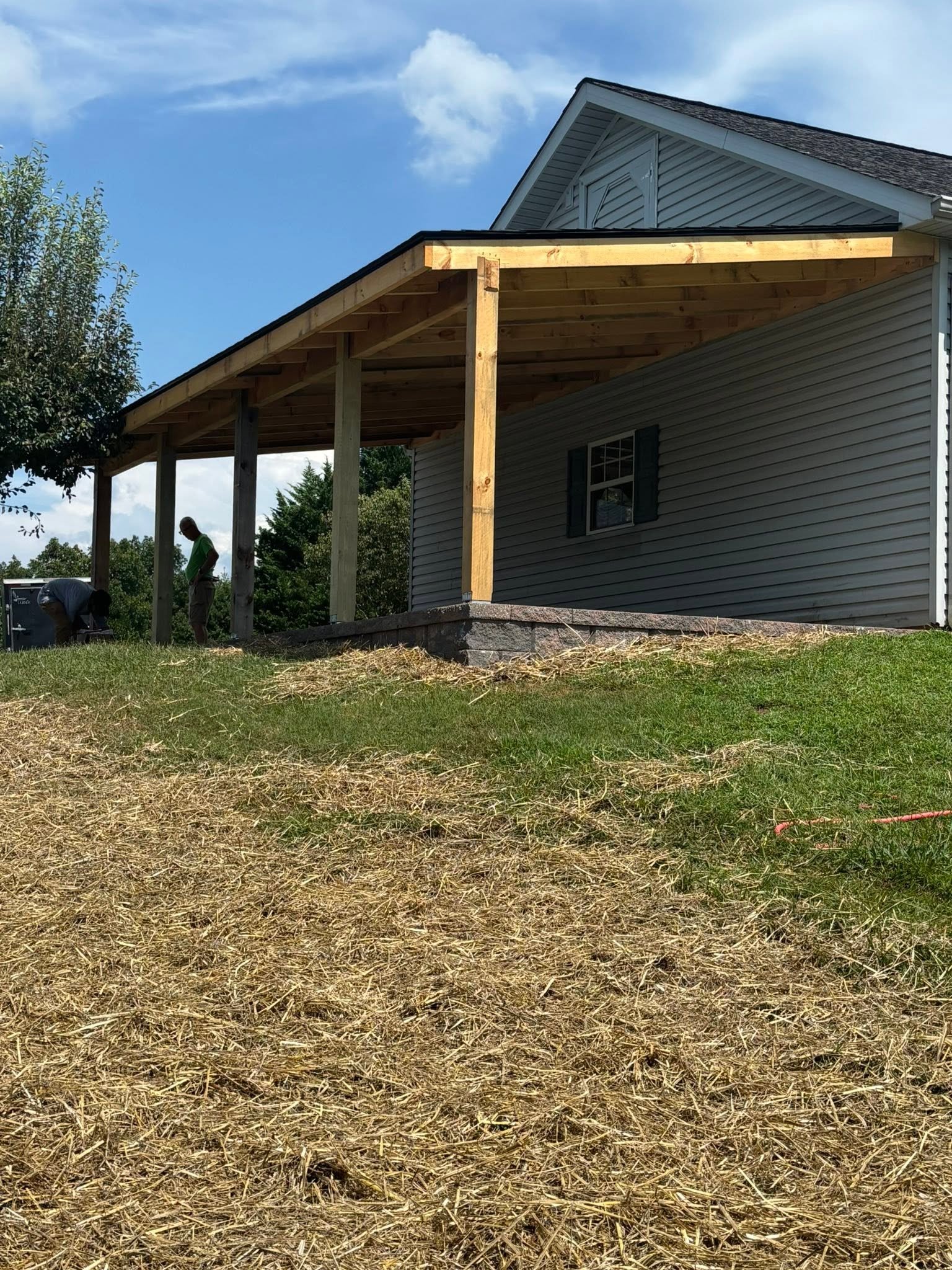 A house under construction with a partially built wooden porch or deck, two workers working nearby, green grass, trees in the background, and a blue sky with some clouds.
