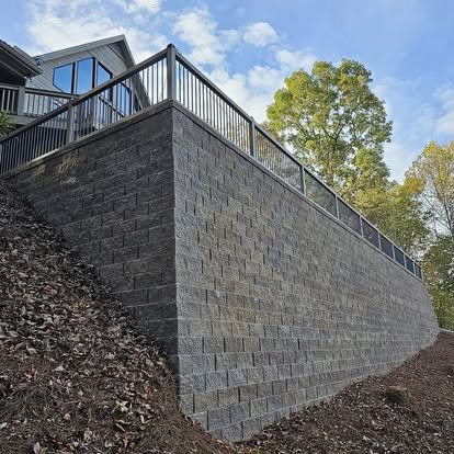 A brick retaining wall supporting a raised patio with a metal railing, with a house and trees in the background under a partly cloudy sky.