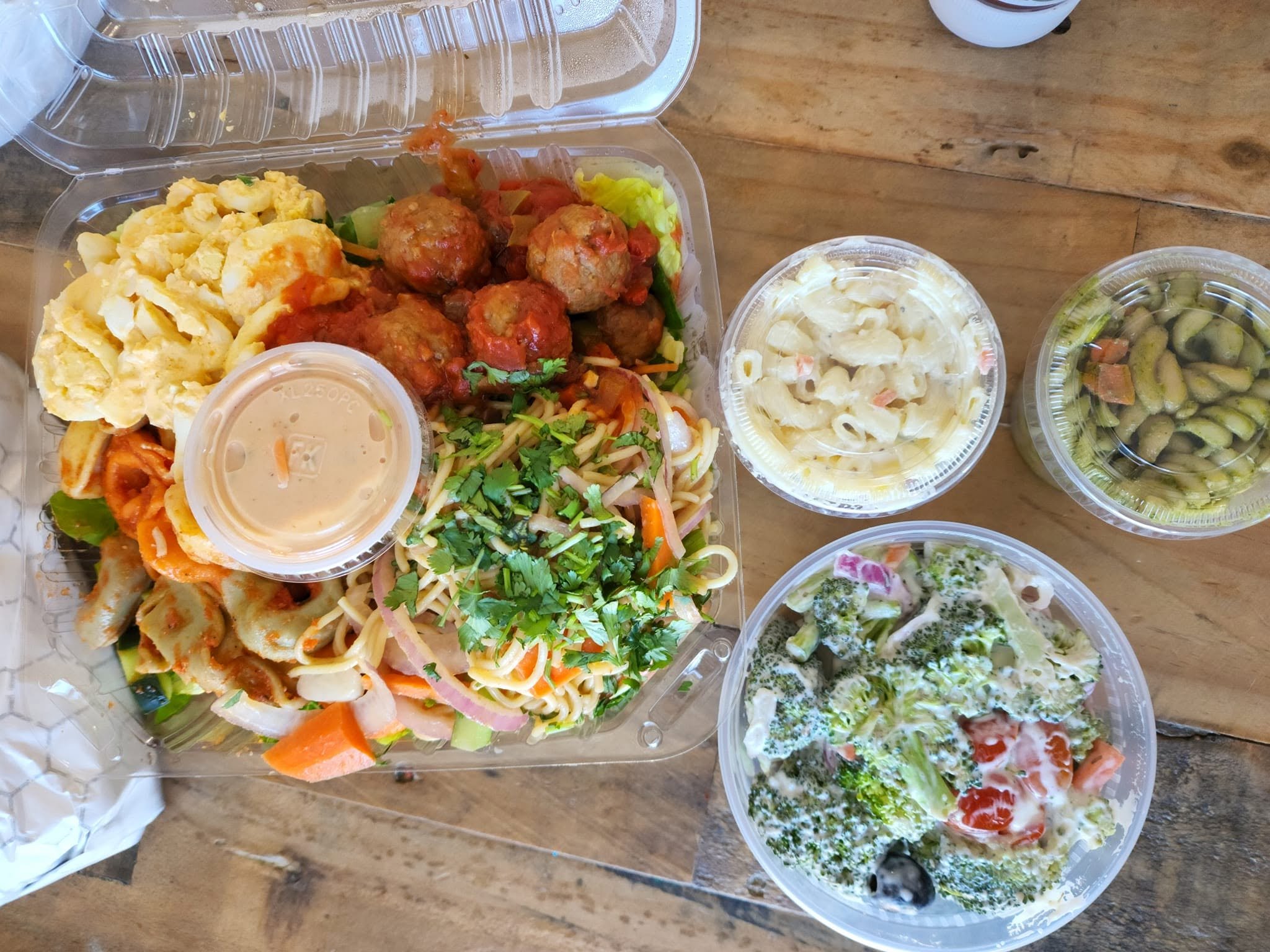 A collection of takeout food containers on a wooden table, including pasta salad, meatballs with sauce, scrambled eggs, and various salads.