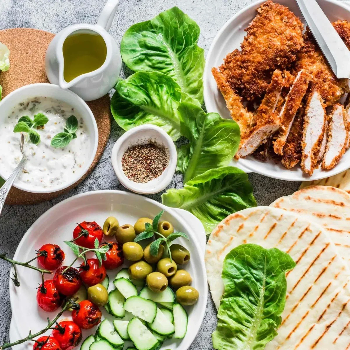 A plate of fried chicken with sliced pieces, surrounded by fresh vegetables including cherry tomatoes, cucumber, lettuce, and green olives, along with small bowls of sauce and spices.