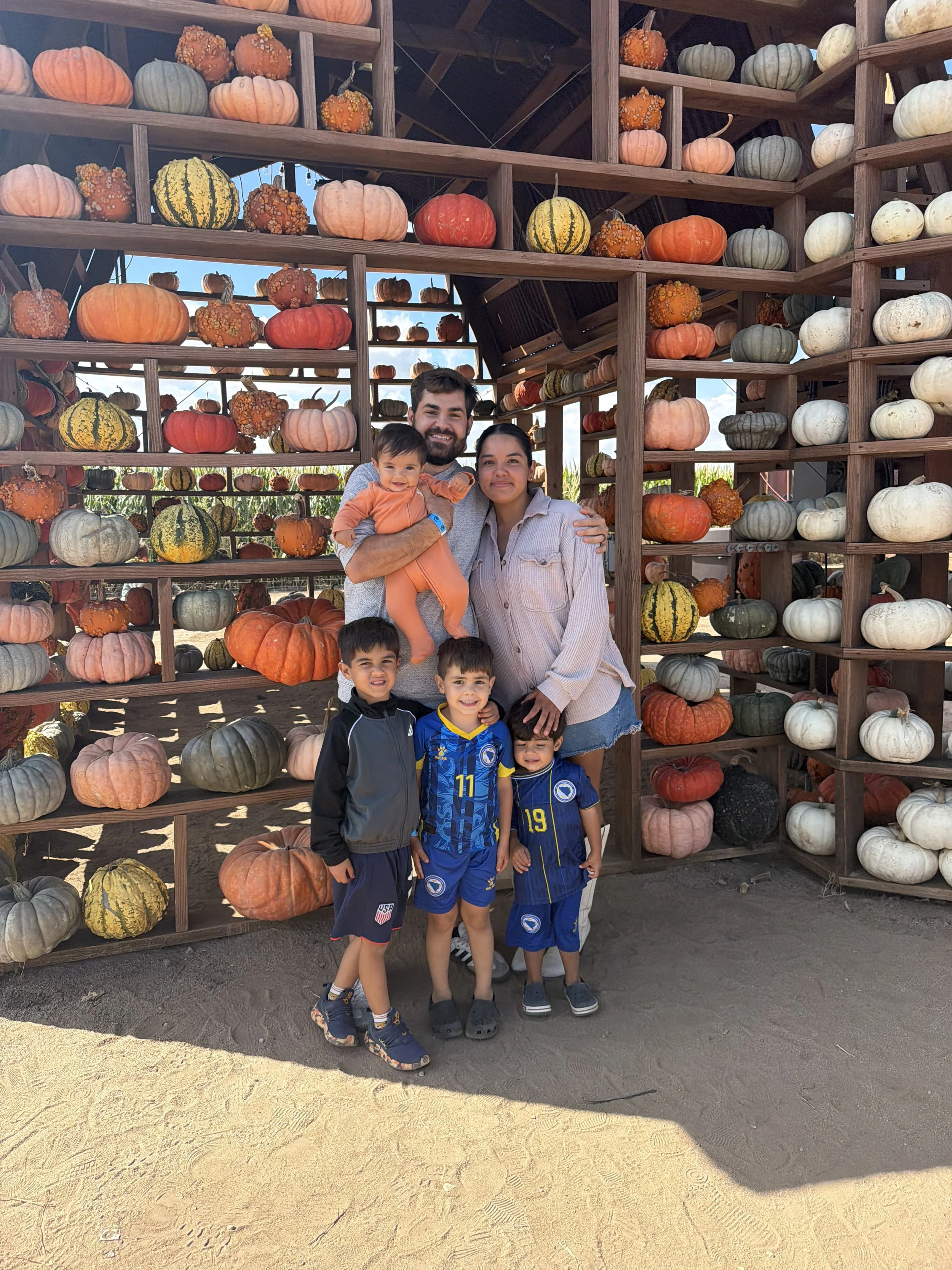 A family of five, two adults and four children, standing in front of a display of pumpkins at a pumpkin patch on a sunny day. The pumpkins are arranged on wooden shelves and are of various colors including orange, white, green, and yellow.