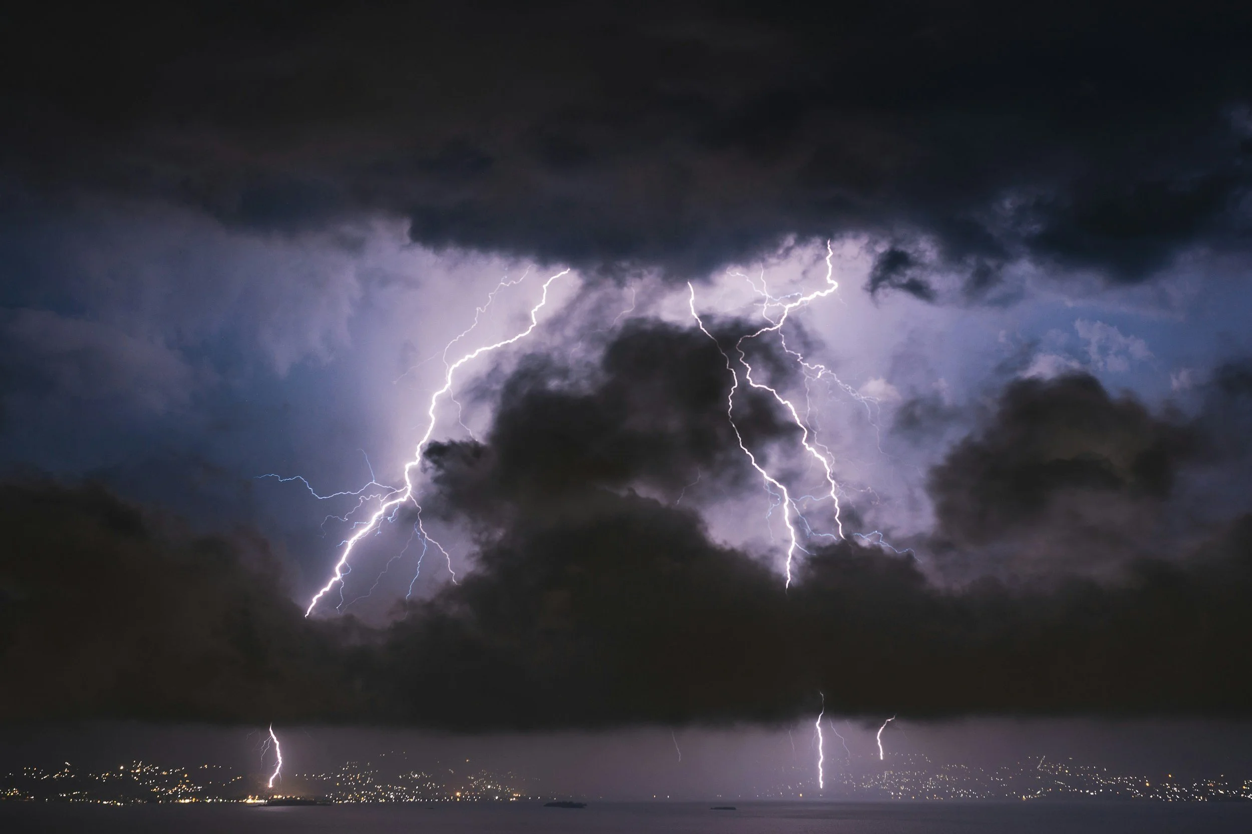 Multiple bolts of lightning striking across a dark cloudy night sky over a body of water with city lights in the distance.