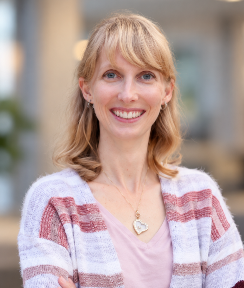 A woman with shoulder-length blonde hair and blue eyes smiling outdoors, wearing a striped cardigan, v-neck pink shirt, and a heart-shaped pendant necklace.