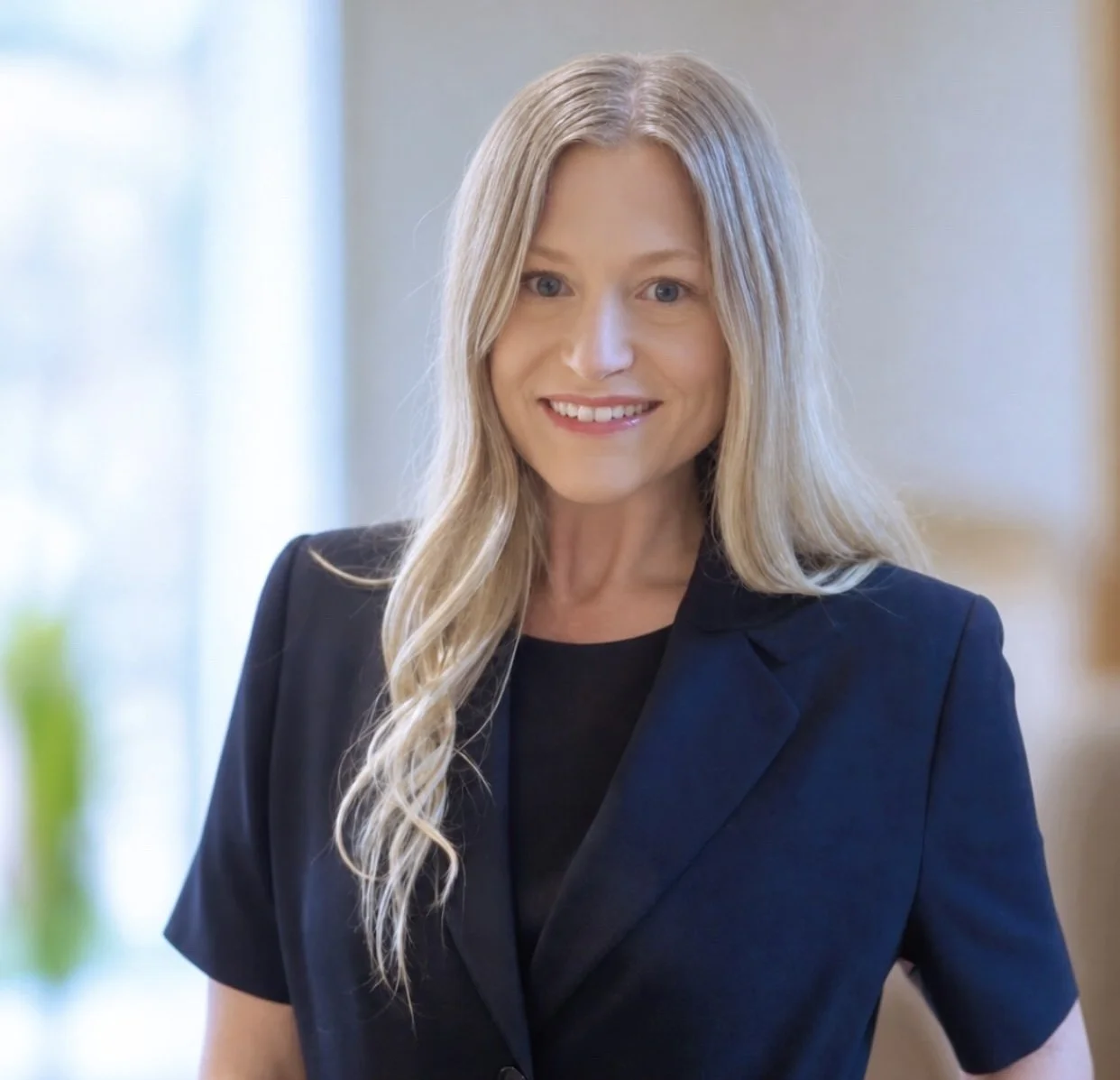 A smiling woman with long blonde hair wearing a black blazer in a well-lit indoor setting.