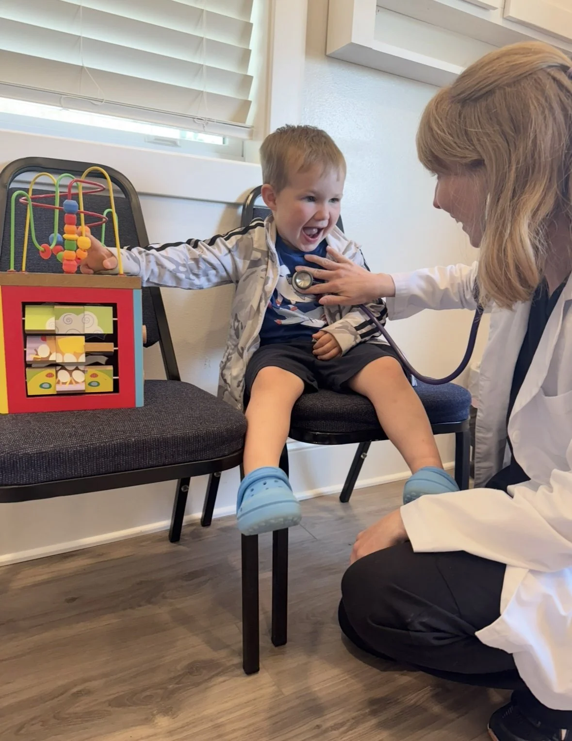 A young boy sitting on a chair, smiling and laughing with a doctor examining him using a stethoscope, with a colorful toy box beside him.