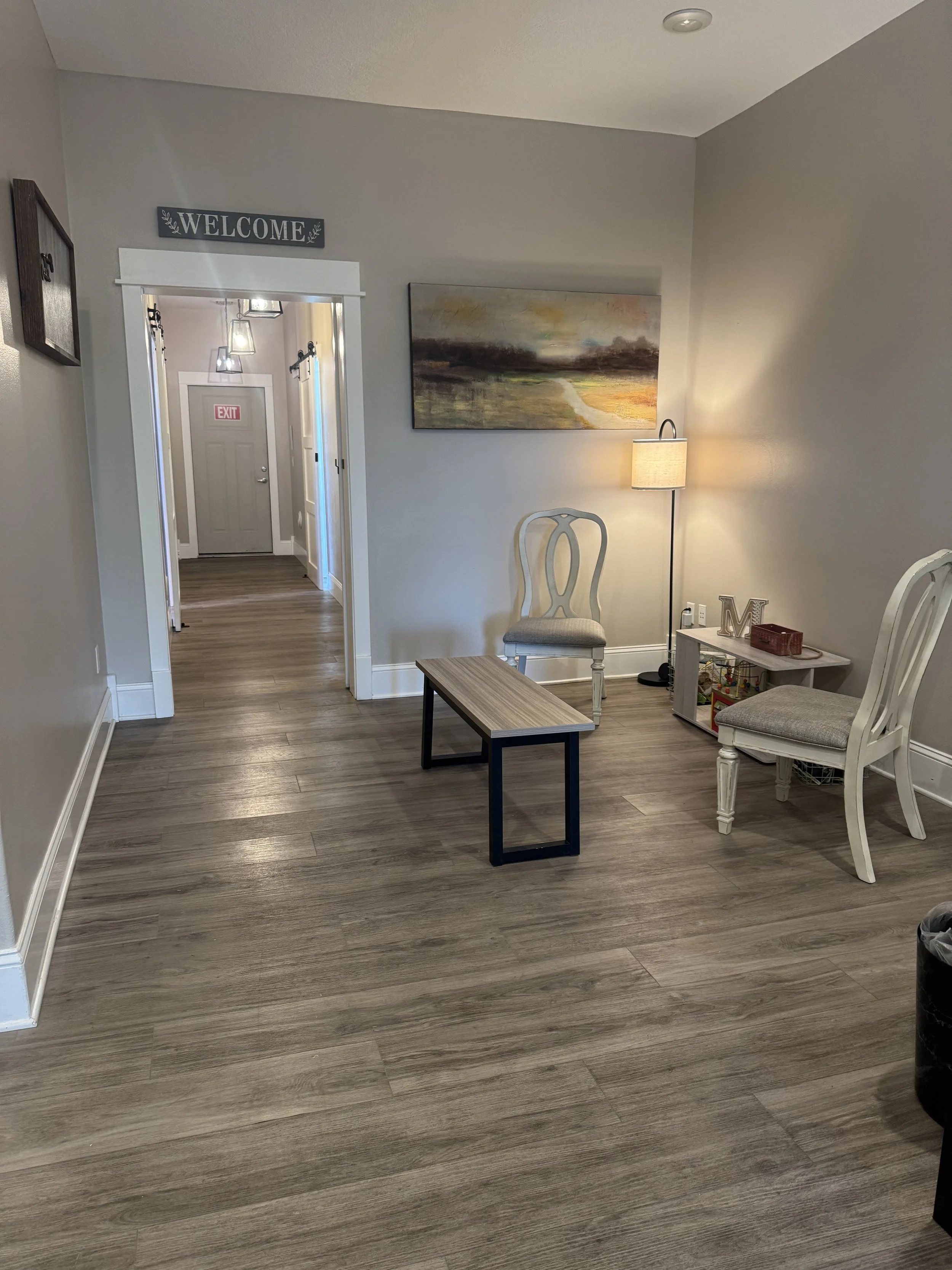 Living room with a wooden floor, white walls, and a white door at the end of a hallway. There are chairs, a small table, a lamp, a landscape painting on the wall, and a sign that reads 'WELCOME' above the doorway.