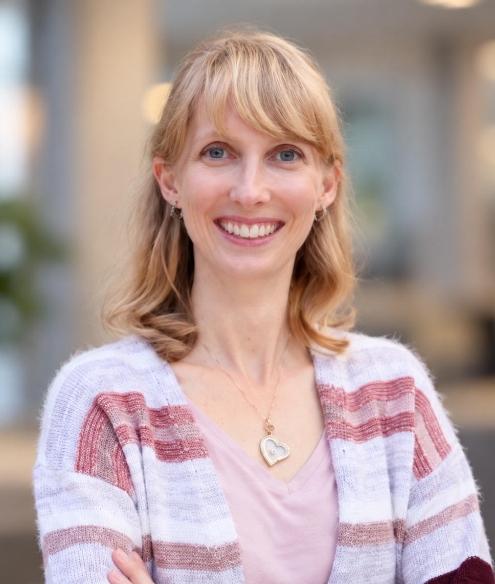 Portrait of a smiling woman with blonde hair, blue eyes, wearing a striped cardigan and a heart-shaped necklace, outdoors with blurred background.