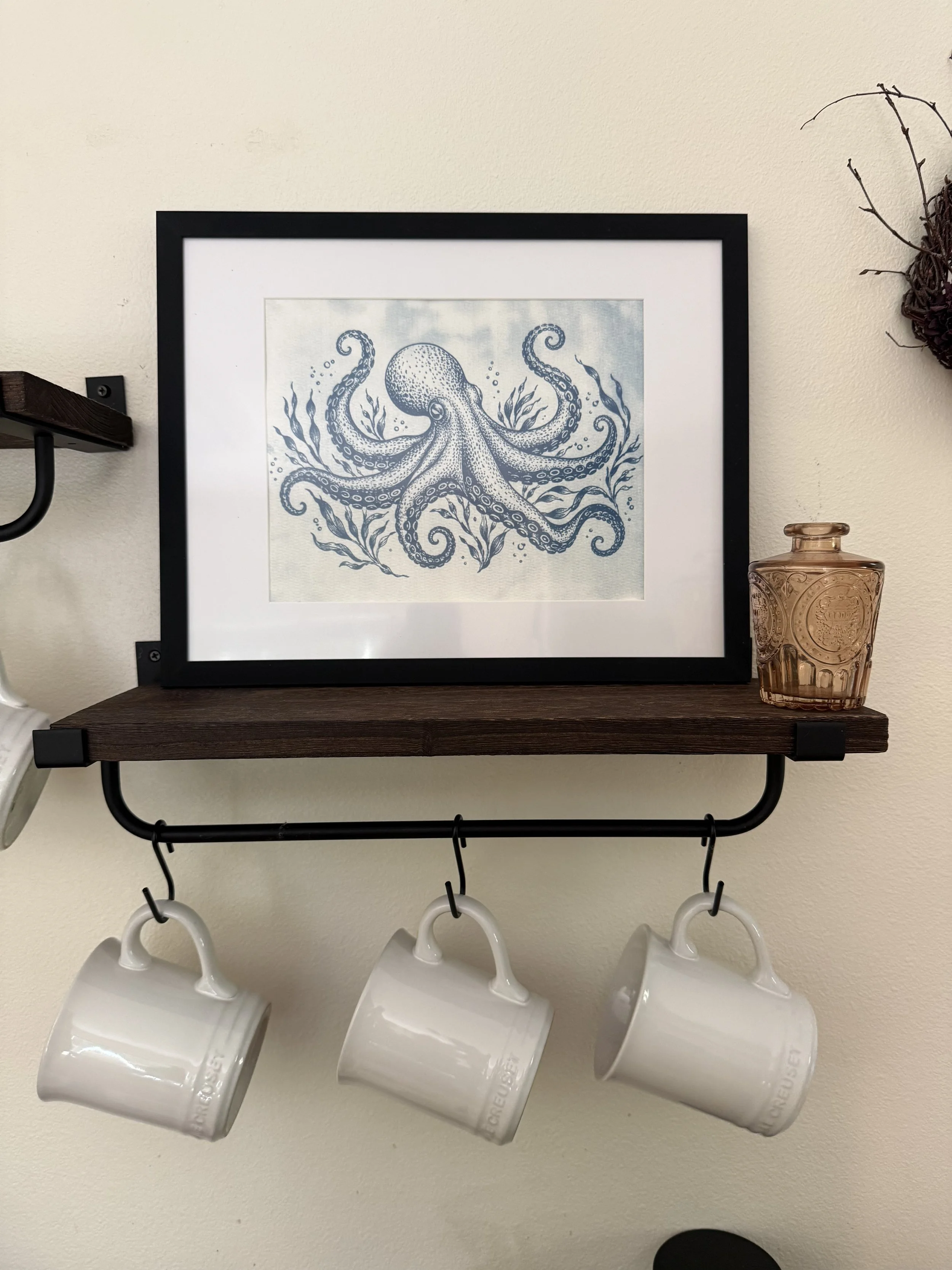Decorative shelf with four white coffee mugs hanging underneath, a framed print of an octopus with seaweed, and a small amber glass bottle on a wooden surface mounted on a wall.