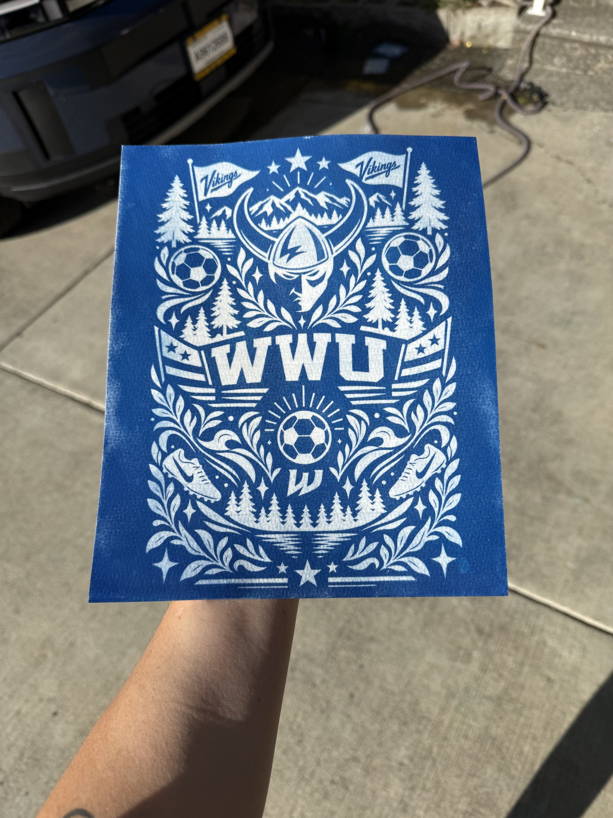 A blue paper with white graphics and the word 'WVU' held in a person's hand, with a parking lot and a car in the background.