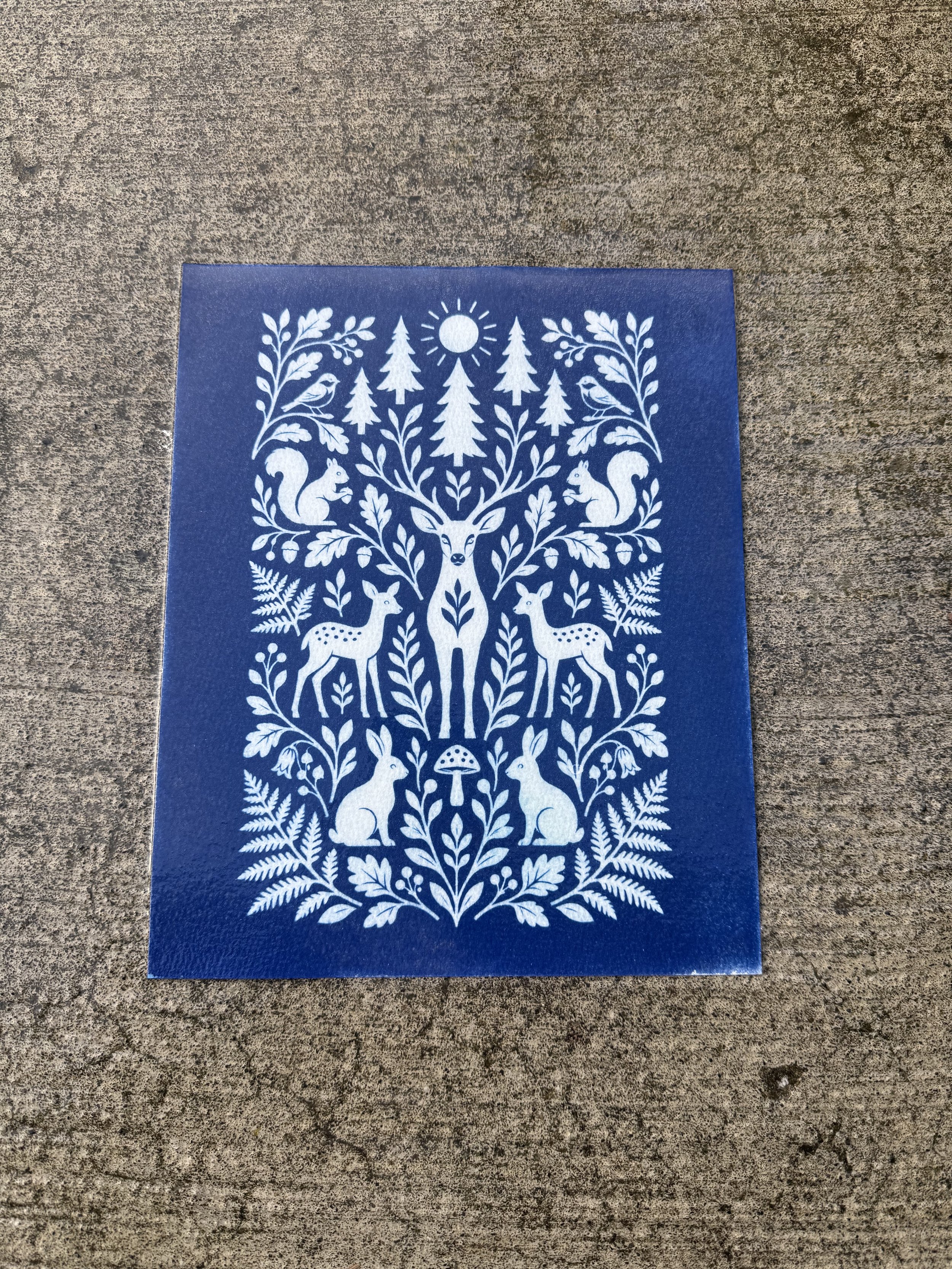 A blue card with white illustrations of woodland animals, trees, and plants arranged symmetrically in a folk art style, featuring a deer with antlers, squirrels, rabbits, birds, mushrooms, and various foliage.