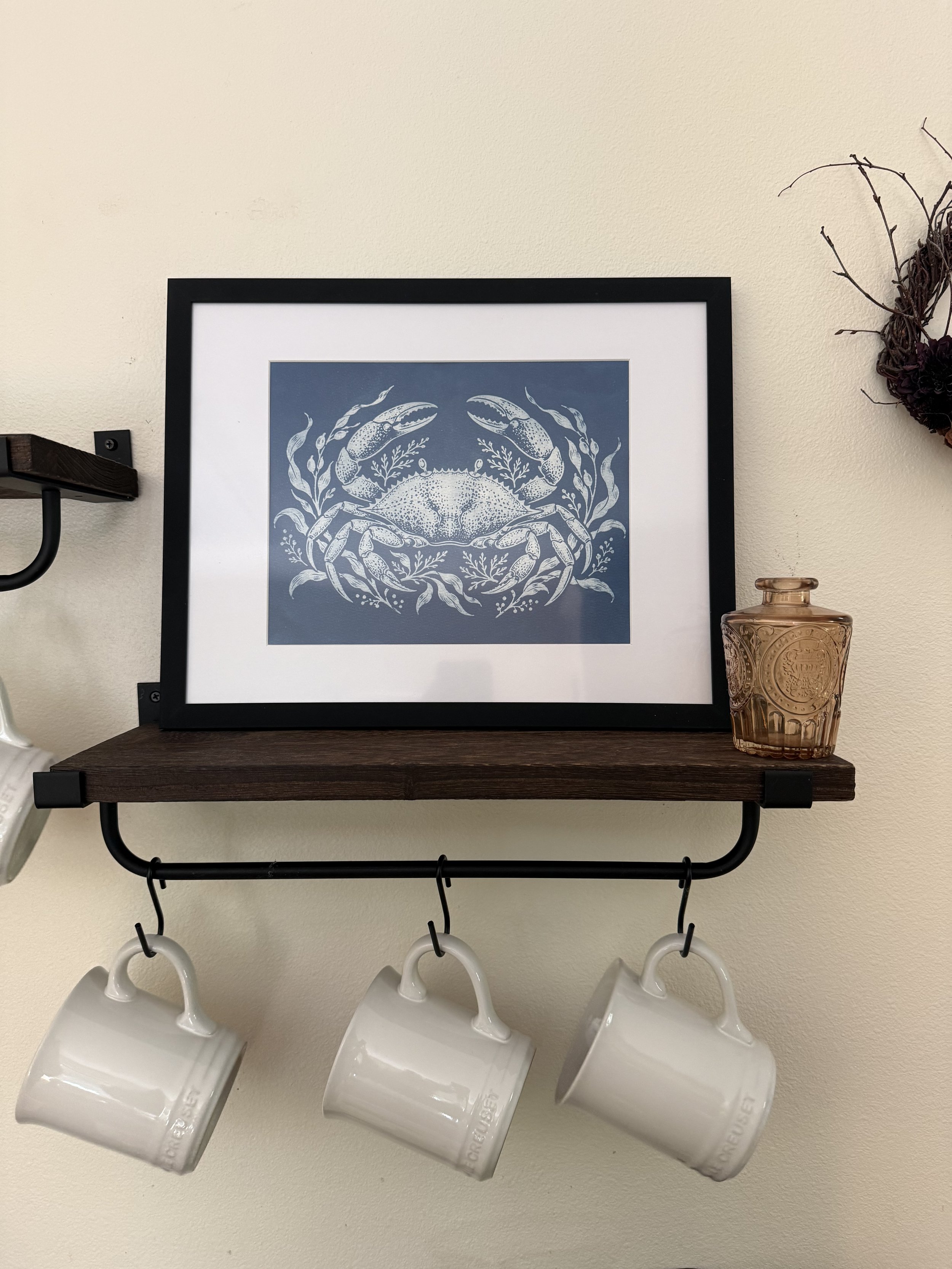 Wall-mounted shelf with four hanging white coffee mugs, a framed crab illustration, a small brown glass jar, and a dried flower arrangement on the right.