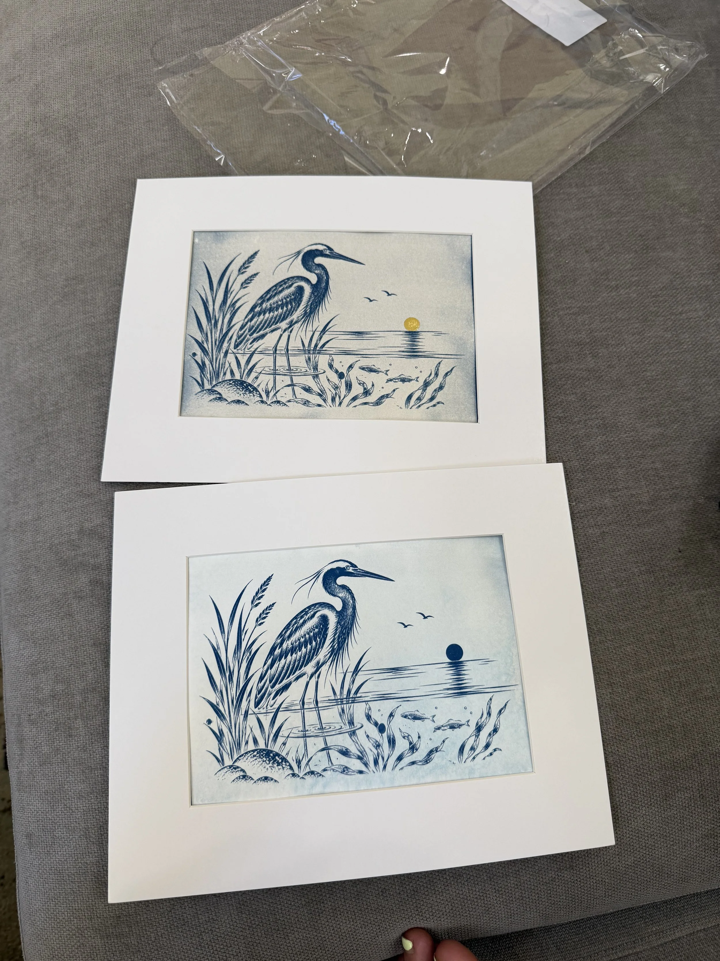 A framed blue and white illustration of a heron standing in a marsh with tall grasses, fish swimming in the water, and a setting sun or moon in the background.