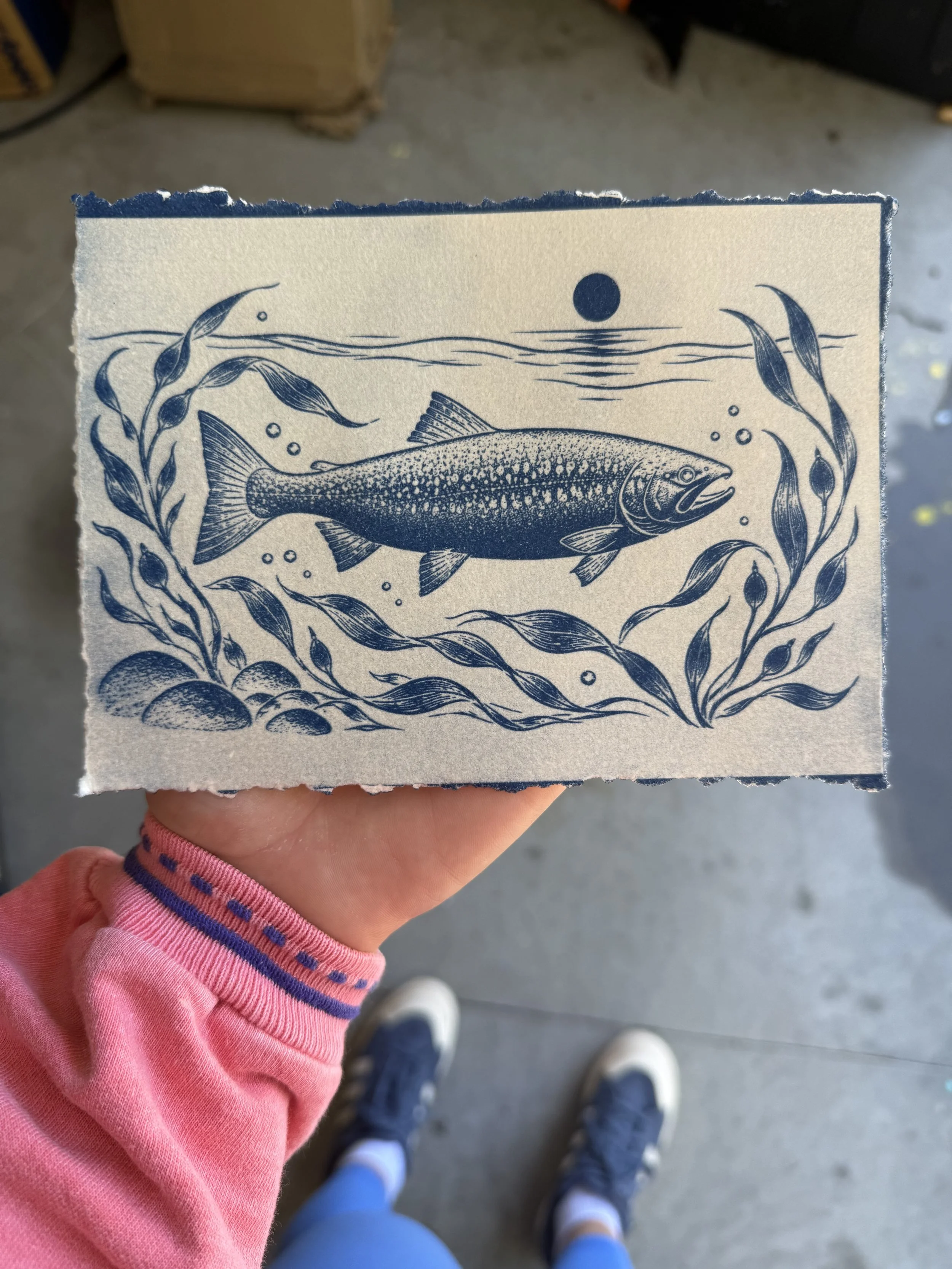 View from above of a person holding a piece of paper with an illustration of a fish and underwater scene, with other people's legs and footwear visible in the background.