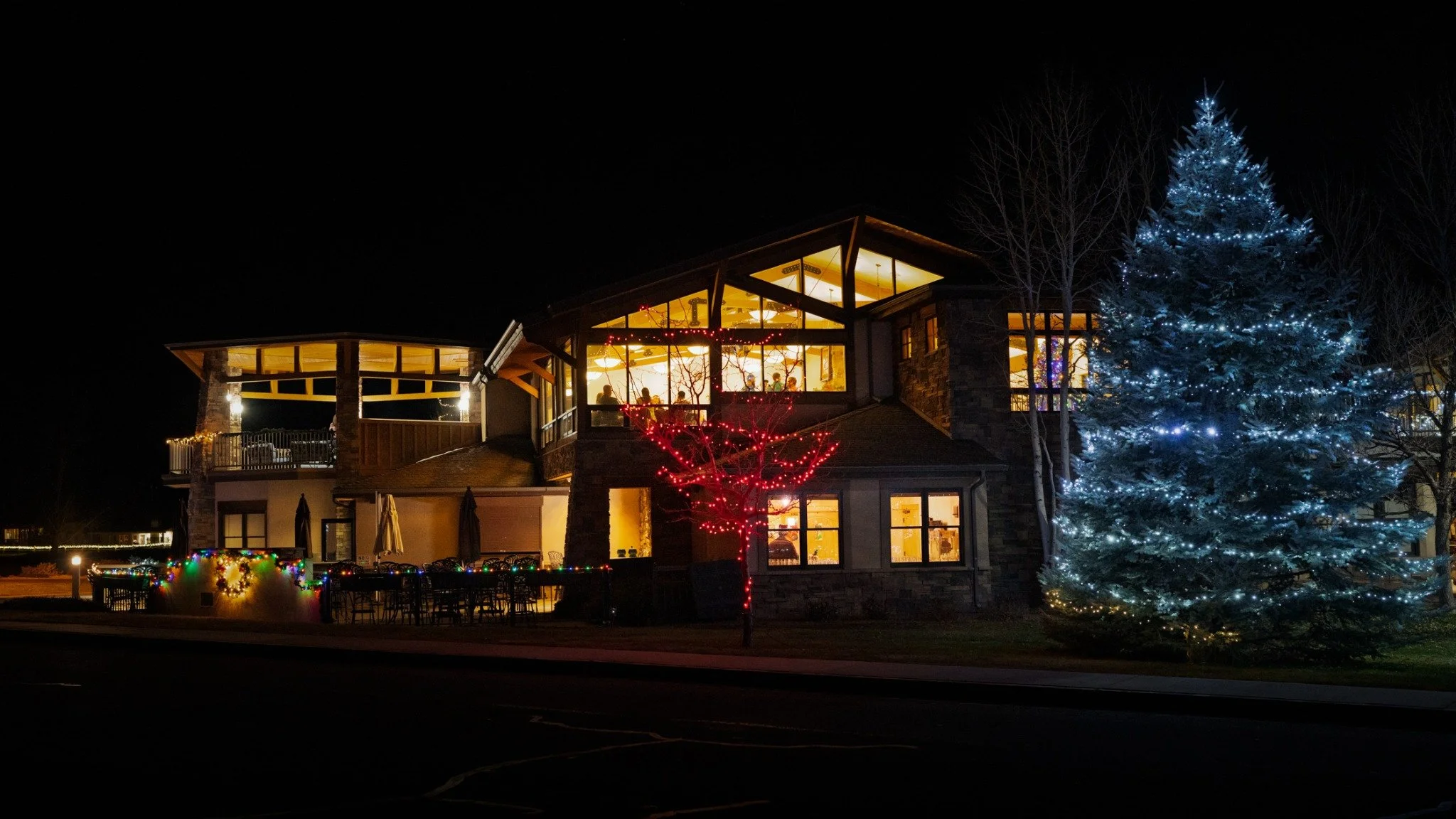 Cobble Creek clubhouse illuminated at night, with red holiday lights and a large evergreen wrapped in white lights.