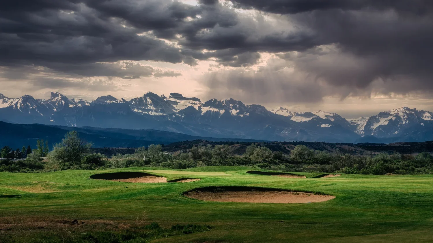 The thirteenth green at Cobble Creek, with bunkers in the foreground and the Cimarron Mountains beneath dramatic clouds.