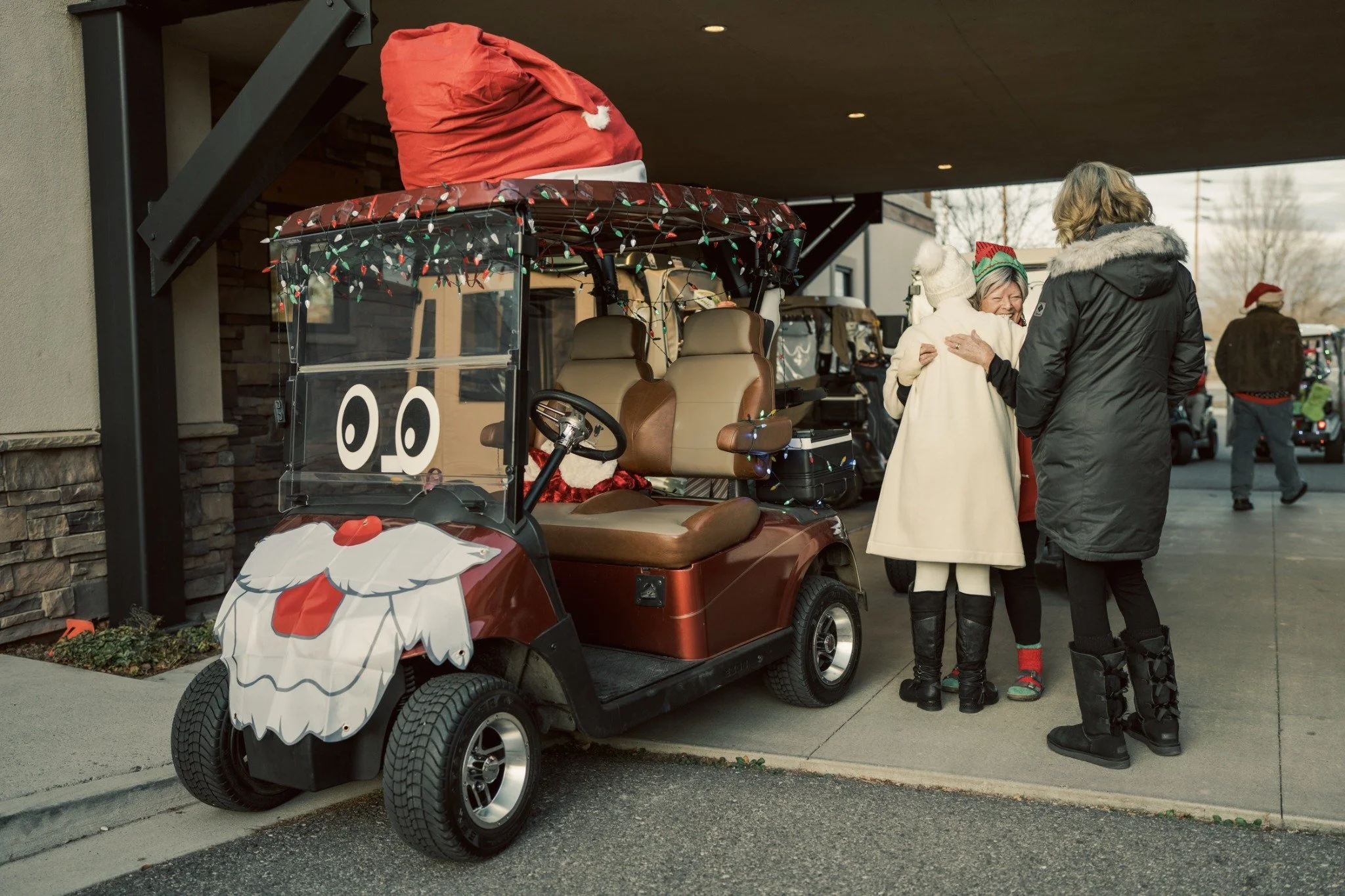 Two people embrace beside a golf cart decorated with an oversized Santa hat and beard at the clubhouse entrance.