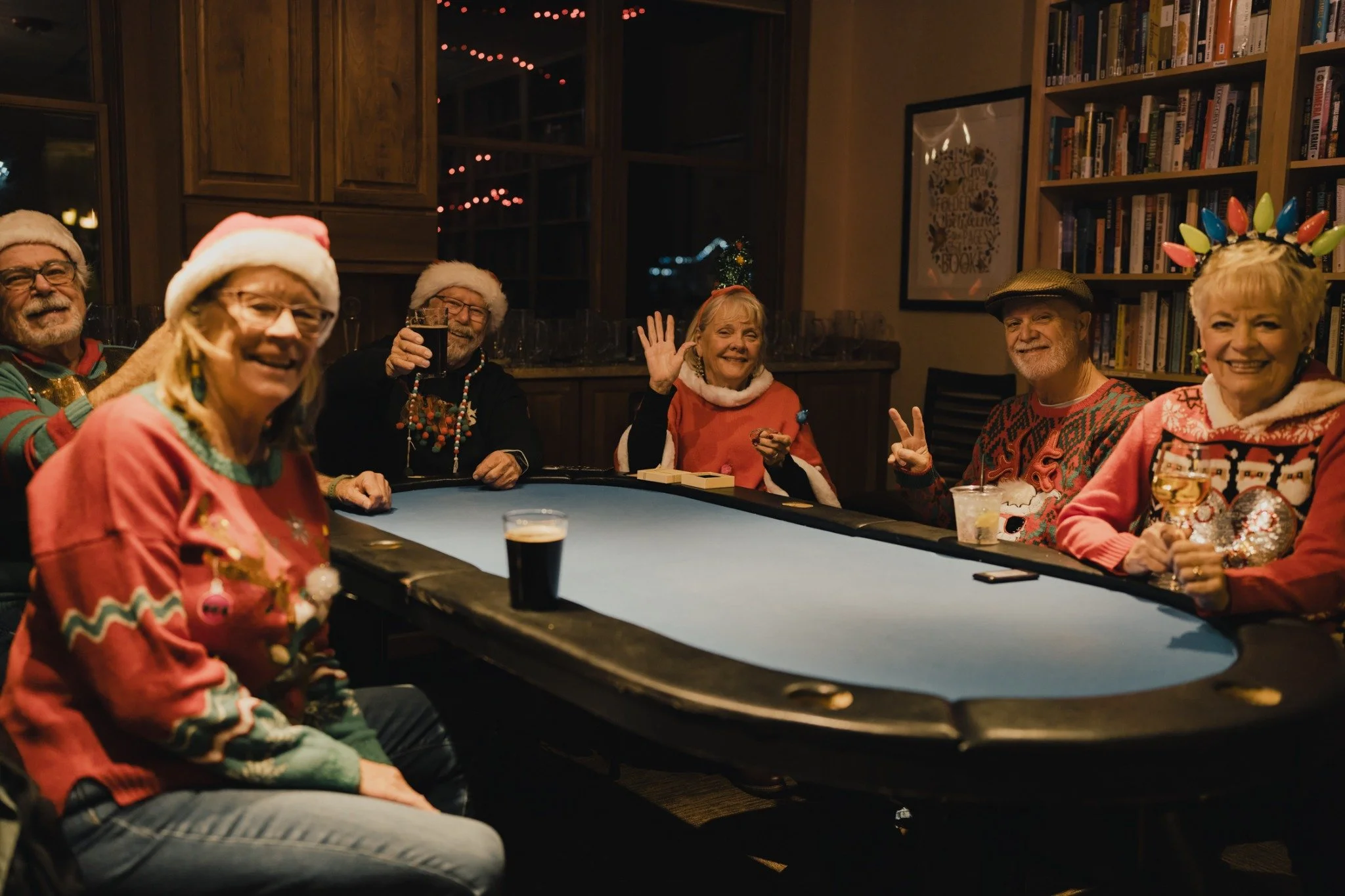 Six people in festive clothing sit around a blue-topped table, with raised glasses and a wave toward the camera.